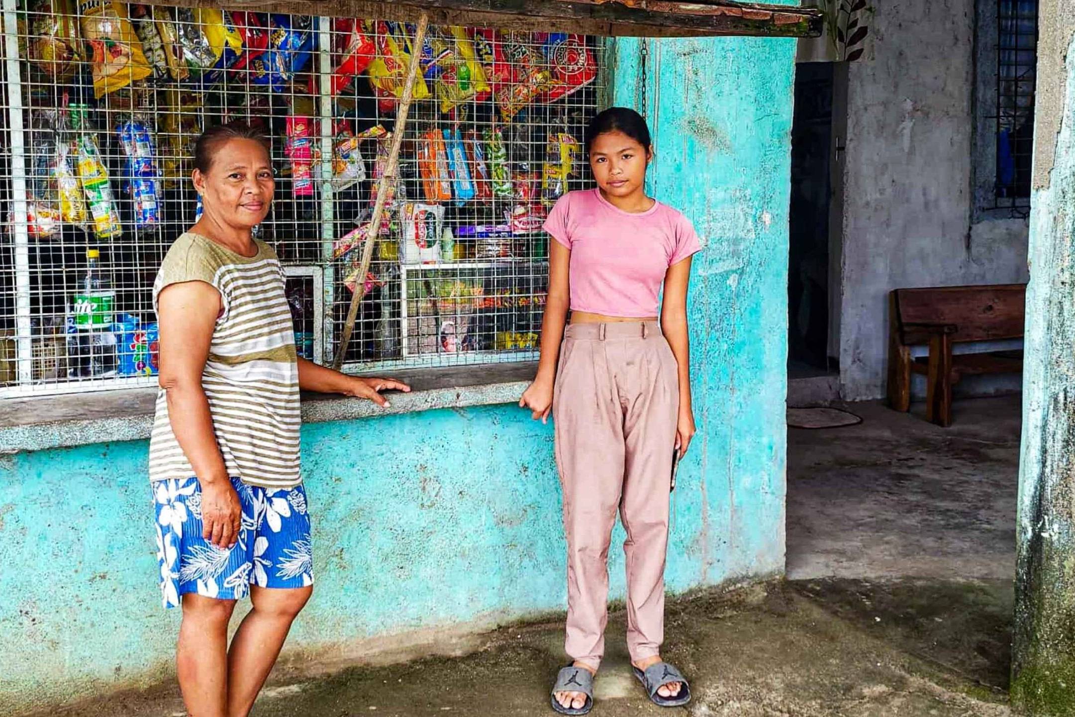Two people stand outside a small shop with teal walls and a metal grille. The woman is in striped shirt and shorts; the girl wears a pink shirt and beige pants. The shop is stocked with snacks, creating a warm community vibe.