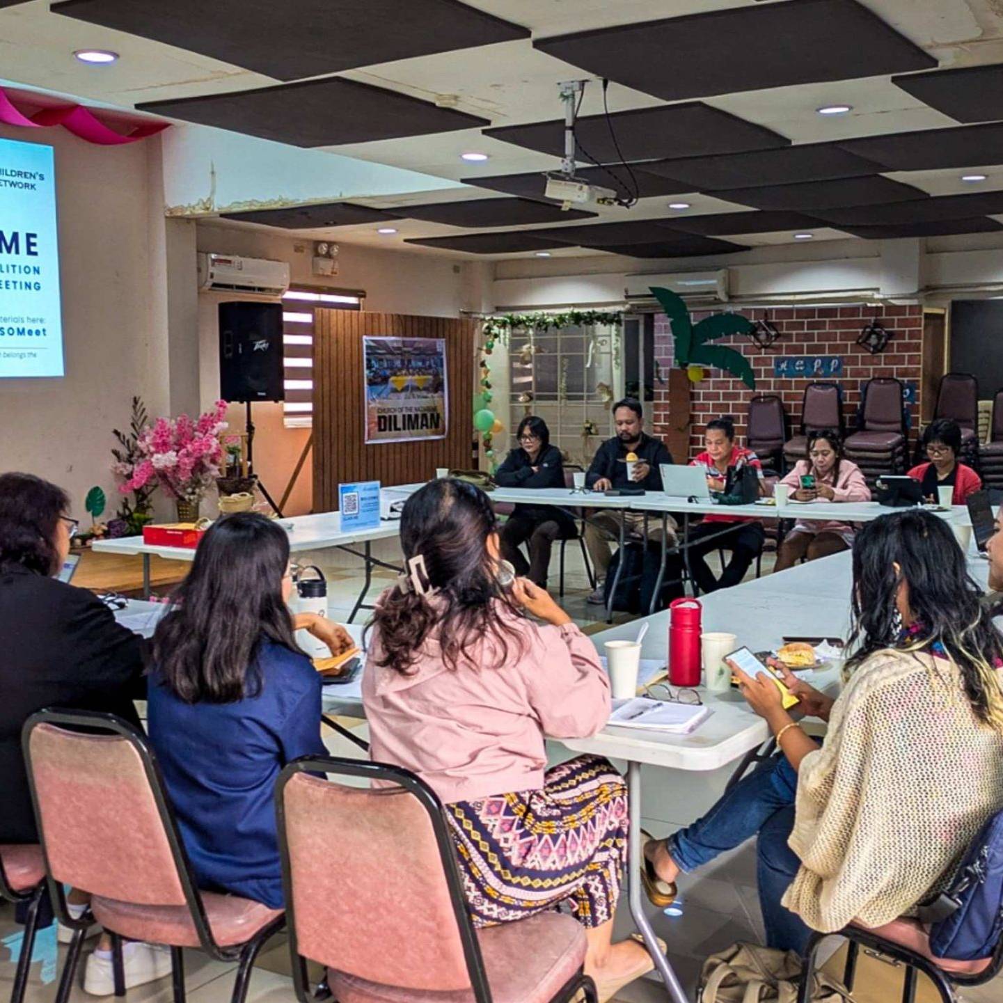 A group of people sits around a large, U-shaped table in a meeting room. The atmosphere is collaborative and engaged.