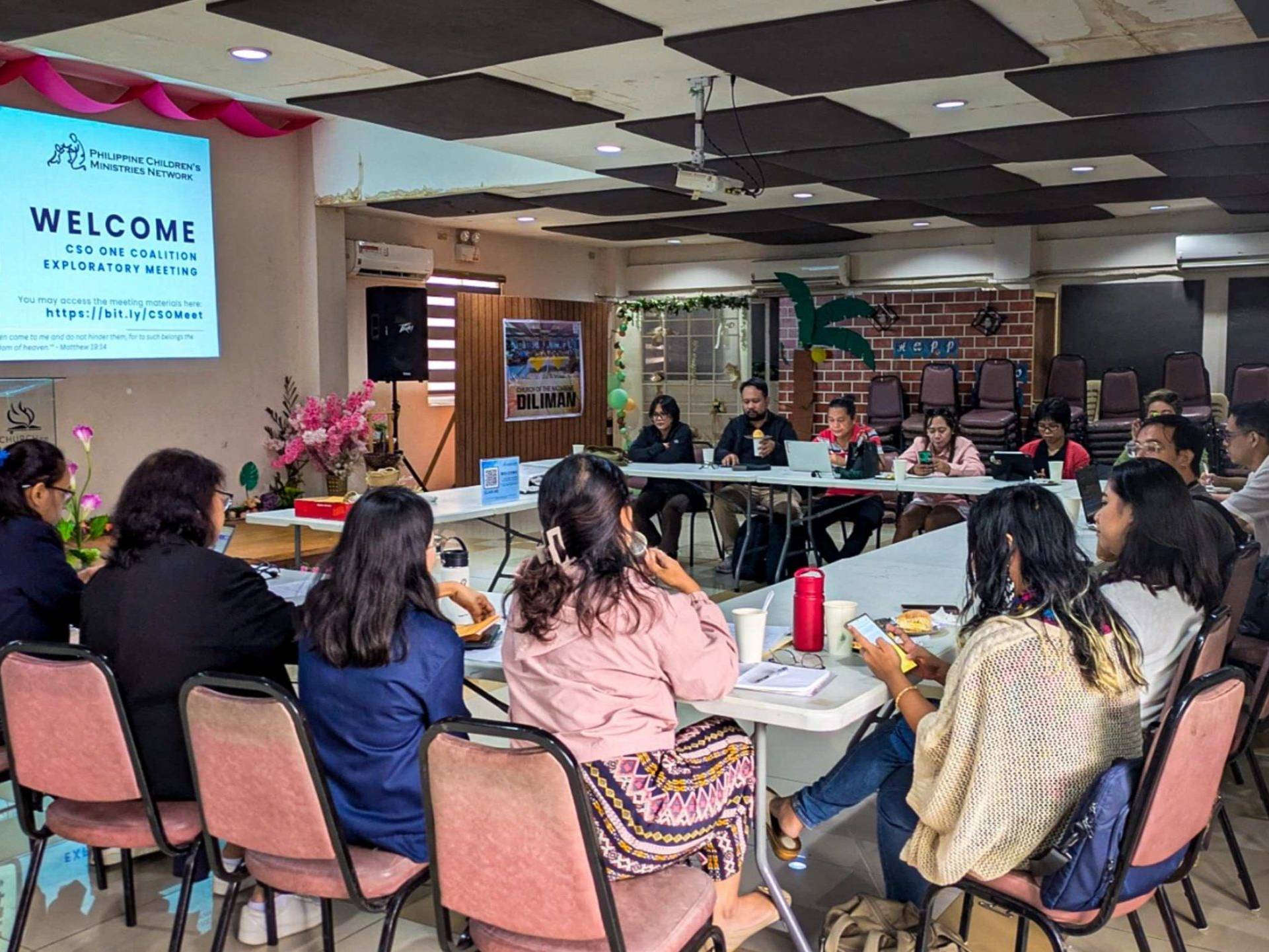 A group of people sits around a large, U-shaped table in a meeting room. The atmosphere is collaborative and engaged.