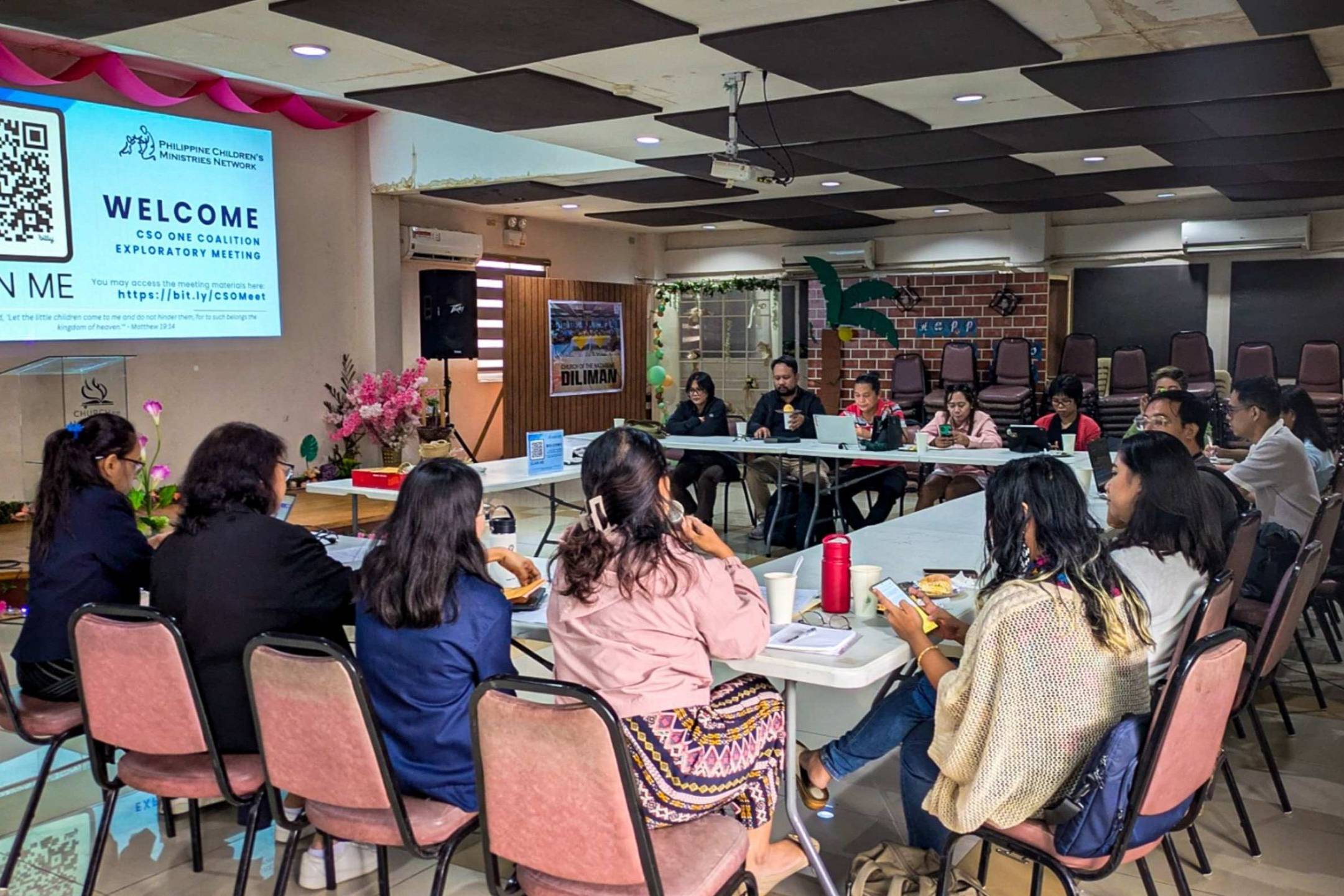 A group of people sits around a large, U-shaped table in a meeting room. The atmosphere is collaborative and engaged.
