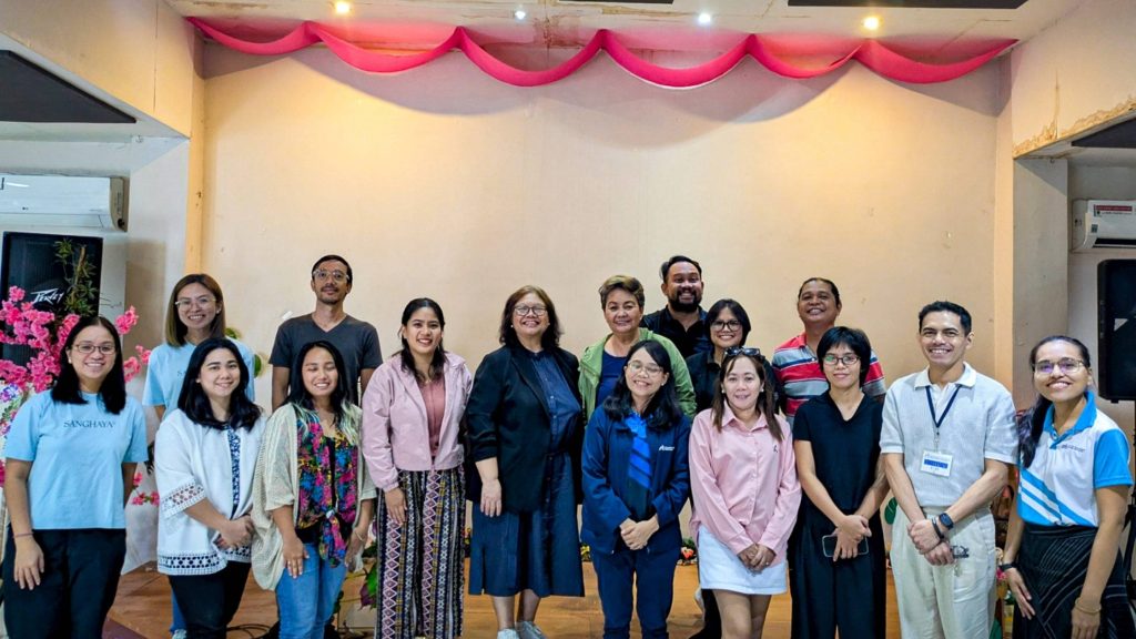 A group of 16 people, mixed genders, smiling in a brightly lit room with pink draped decorations. A joyful, celebratory atmosphere.