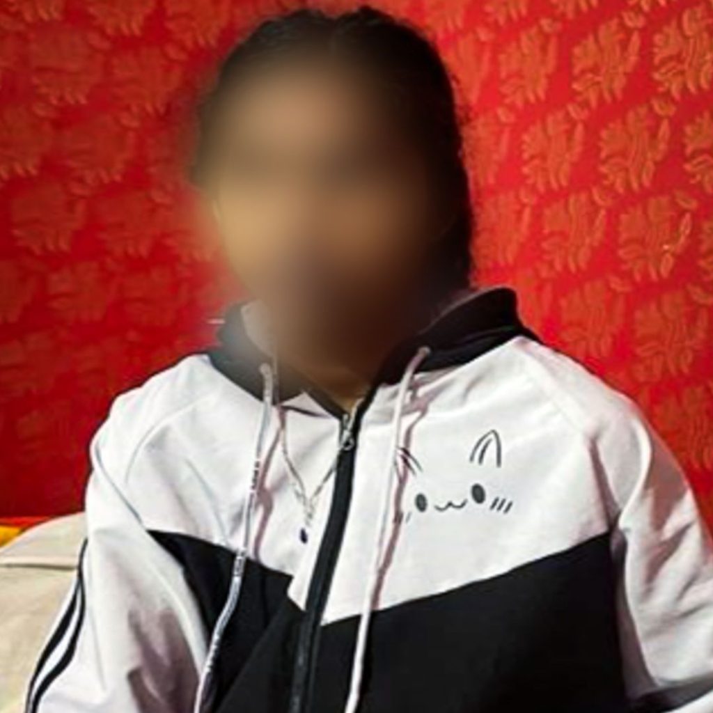 A person with a blurred face wears a black and white hoodie with a cute cat face design. They sit in front of a red patterned wall, creating a relaxed atmosphere.