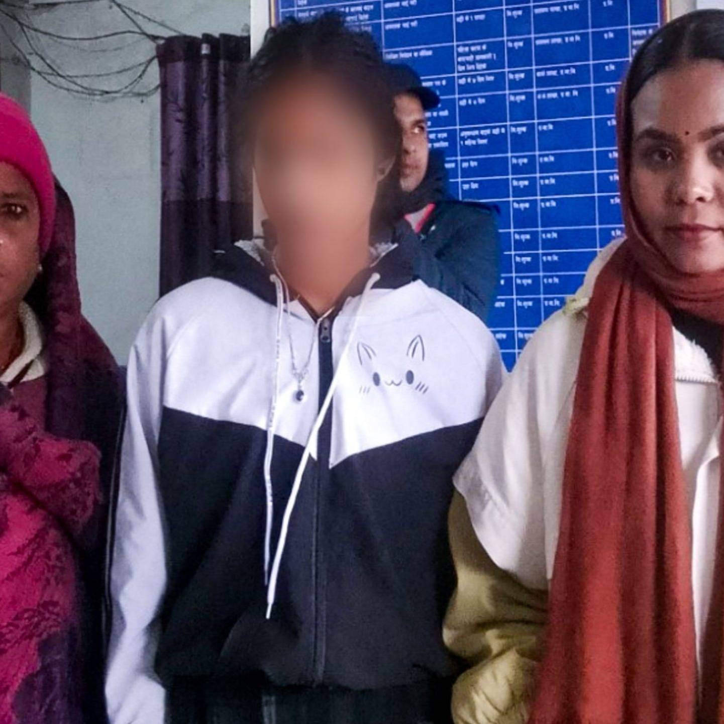 Three people stand indoors, wearing winter clothing. The person in the center has their face blurred. A bulletin board with text is visible in the background.