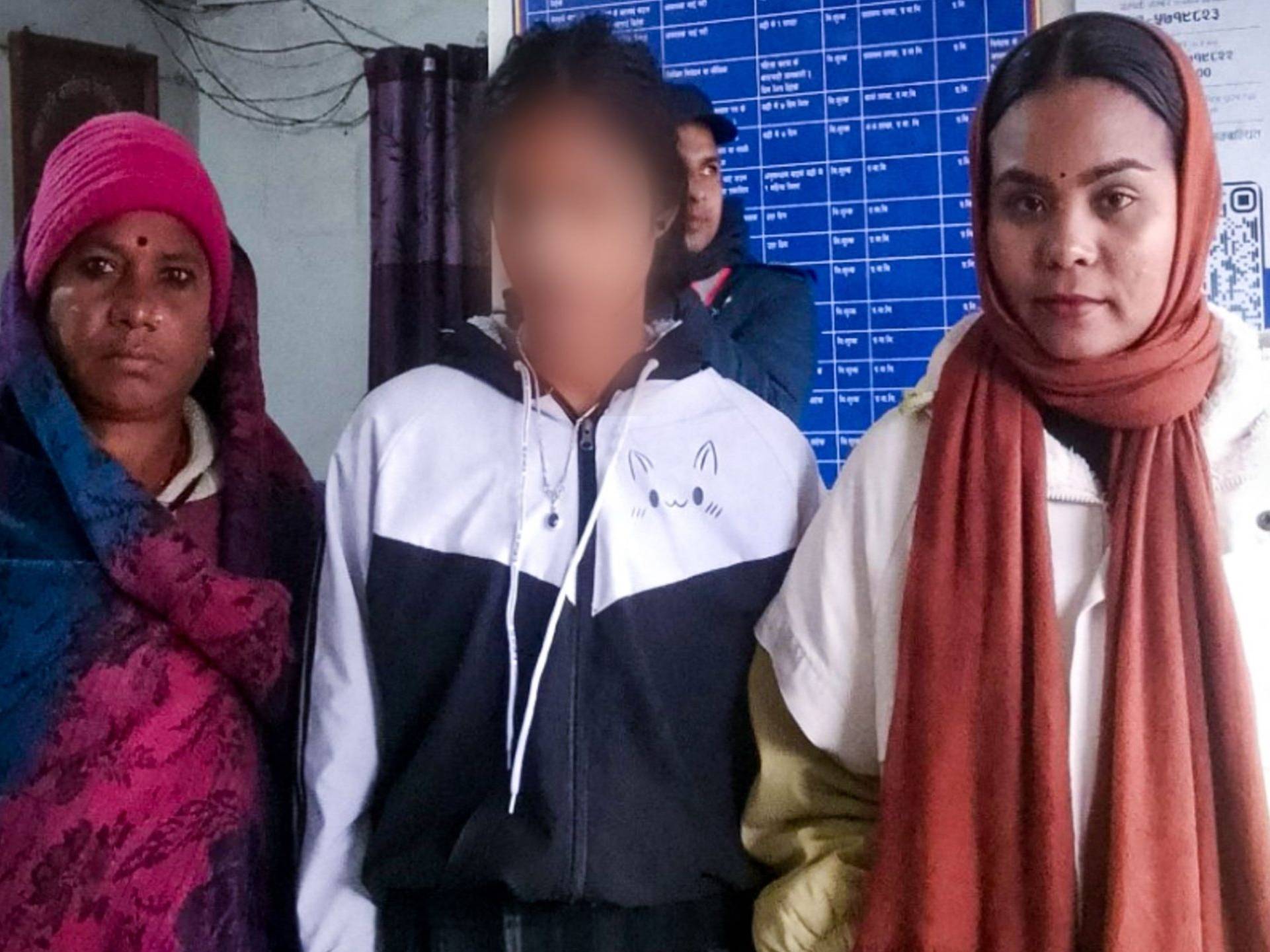 Three people stand indoors, wearing winter clothing. The person in the center has their face blurred. A bulletin board with text is visible in the background.