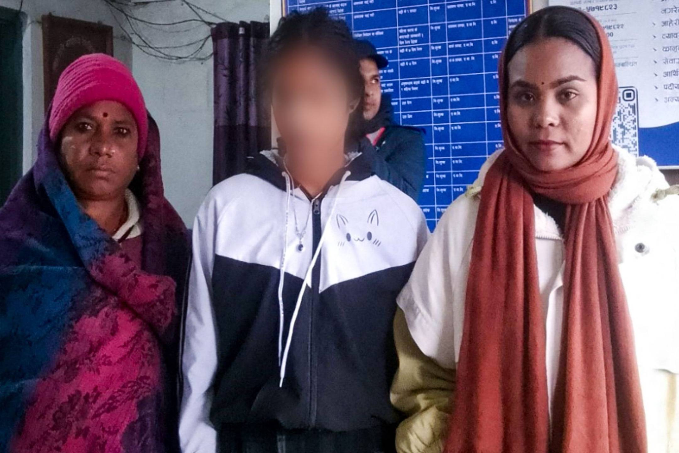 Three people stand indoors, wearing winter clothing. The person in the center has their face blurred. A bulletin board with text is visible in the background.