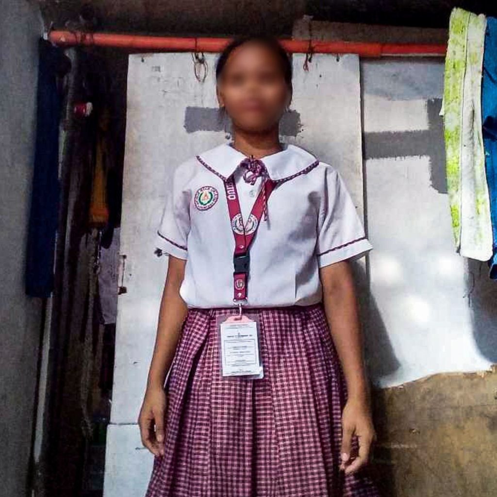 A student in a school uniform stands indoors, with a blurred face for privacy. She wears a white blouse and checkered skirt, conveying a formal, neutral tone.