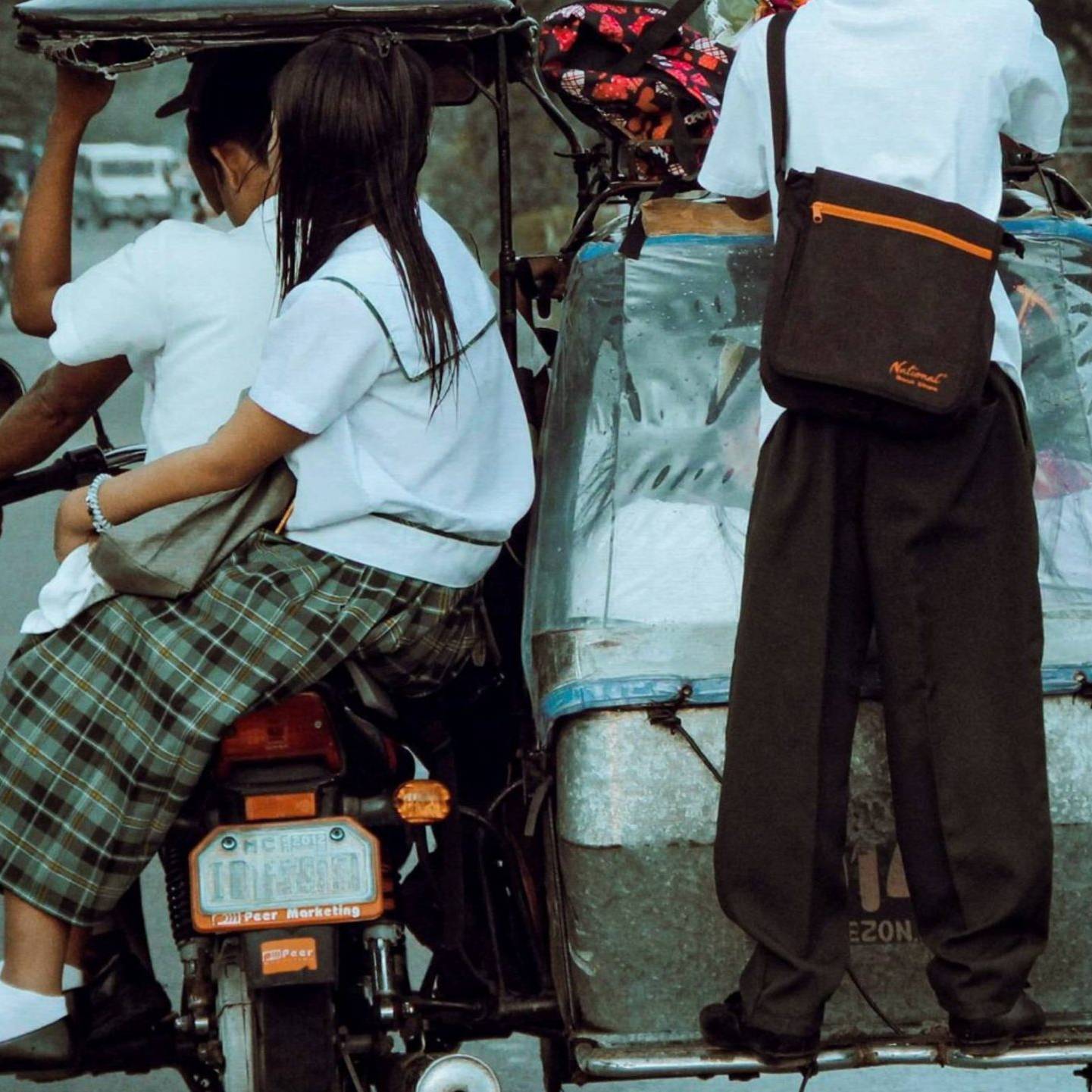 Three students in uniforms ride a tricycle on a rural road. One stands at the back, while two sit. The scene conveys casual, everyday life.