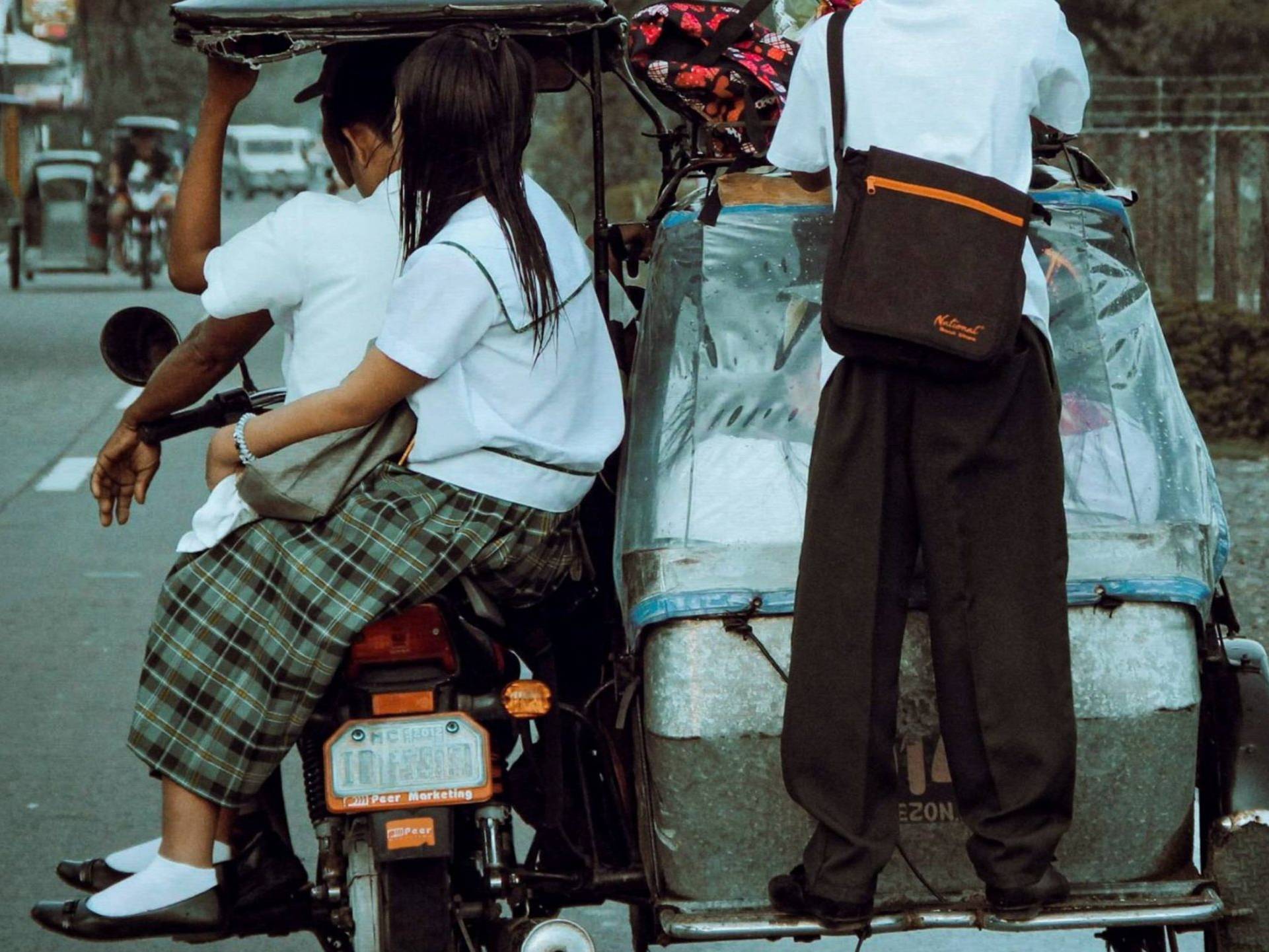 Three students in uniforms ride a tricycle on a rural road. One stands at the back, while two sit. The scene conveys casual, everyday life.