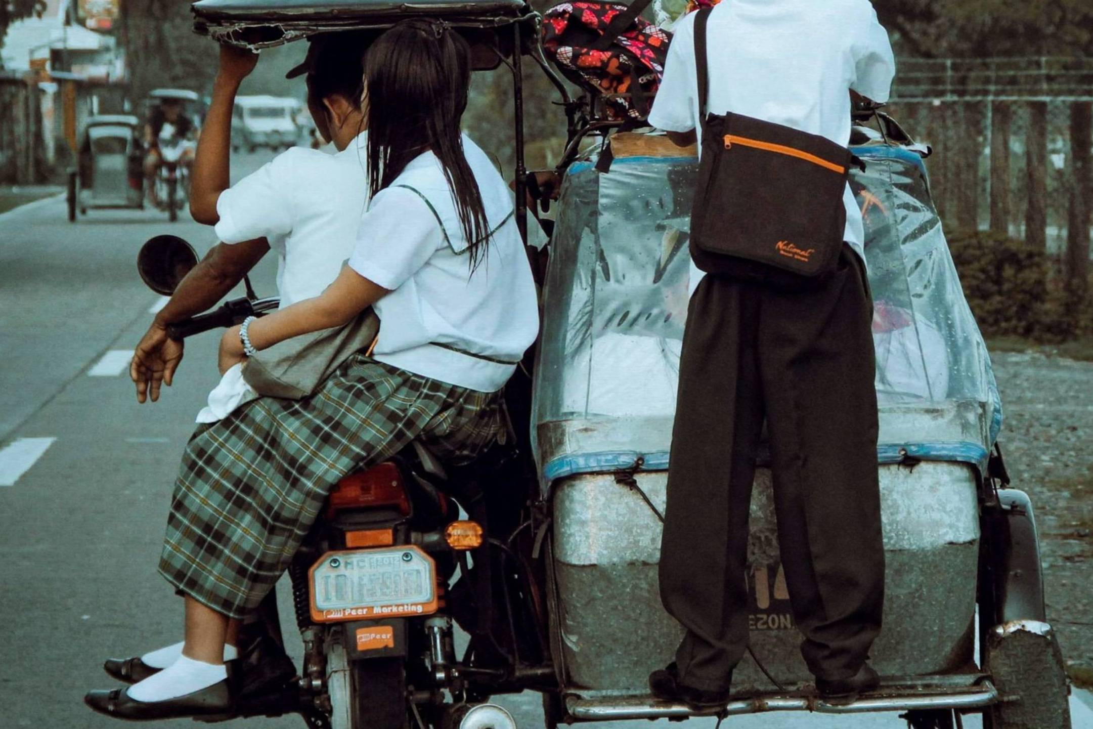 Three students in uniforms ride a tricycle on a rural road. One stands at the back, while two sit. The scene conveys casual, everyday life.