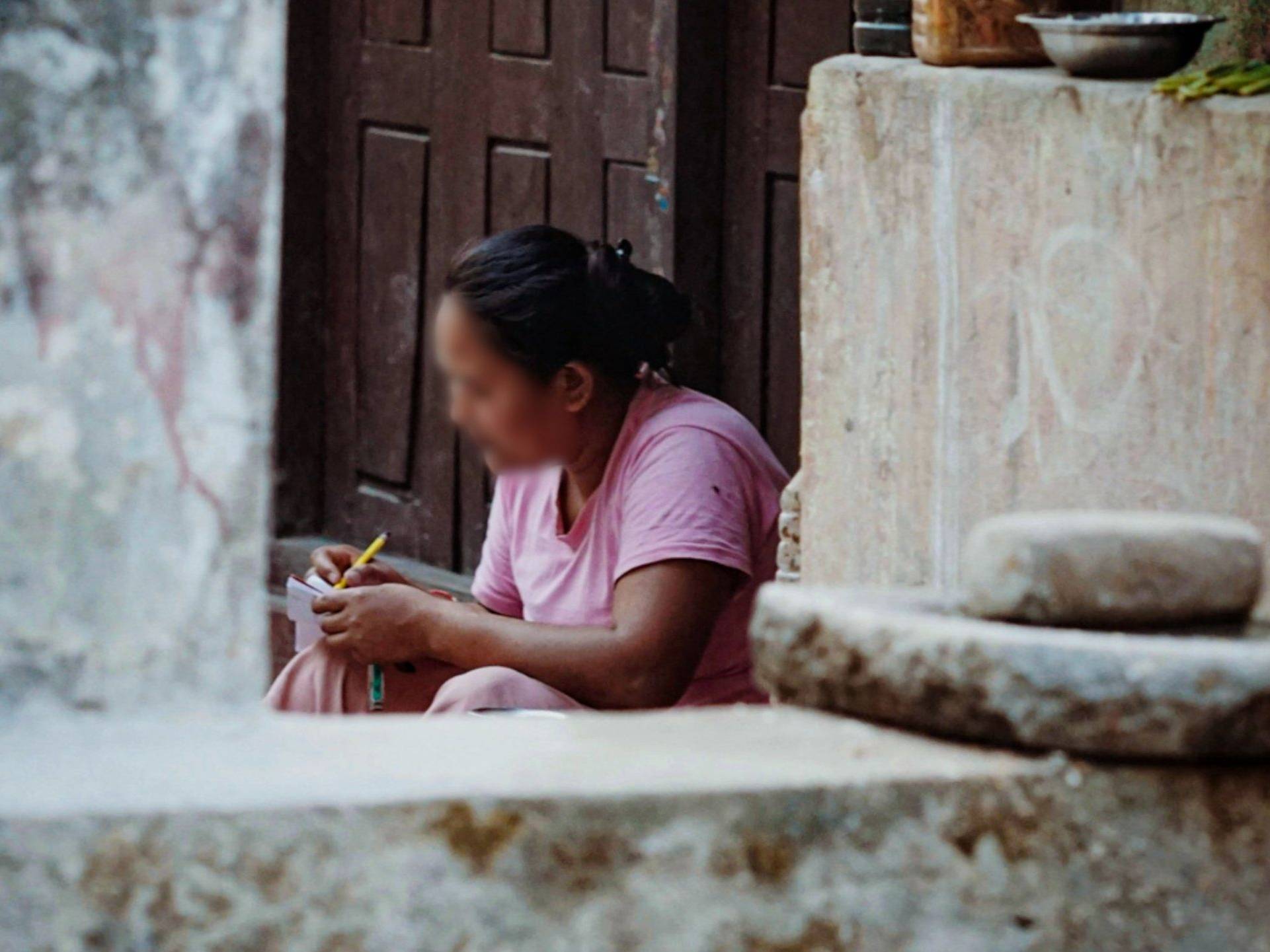 A woman in a pink sari sits on a stone step, writing in a notebook. The scene conveys focus and simplicity, with rustic surroundings including a stone wall.