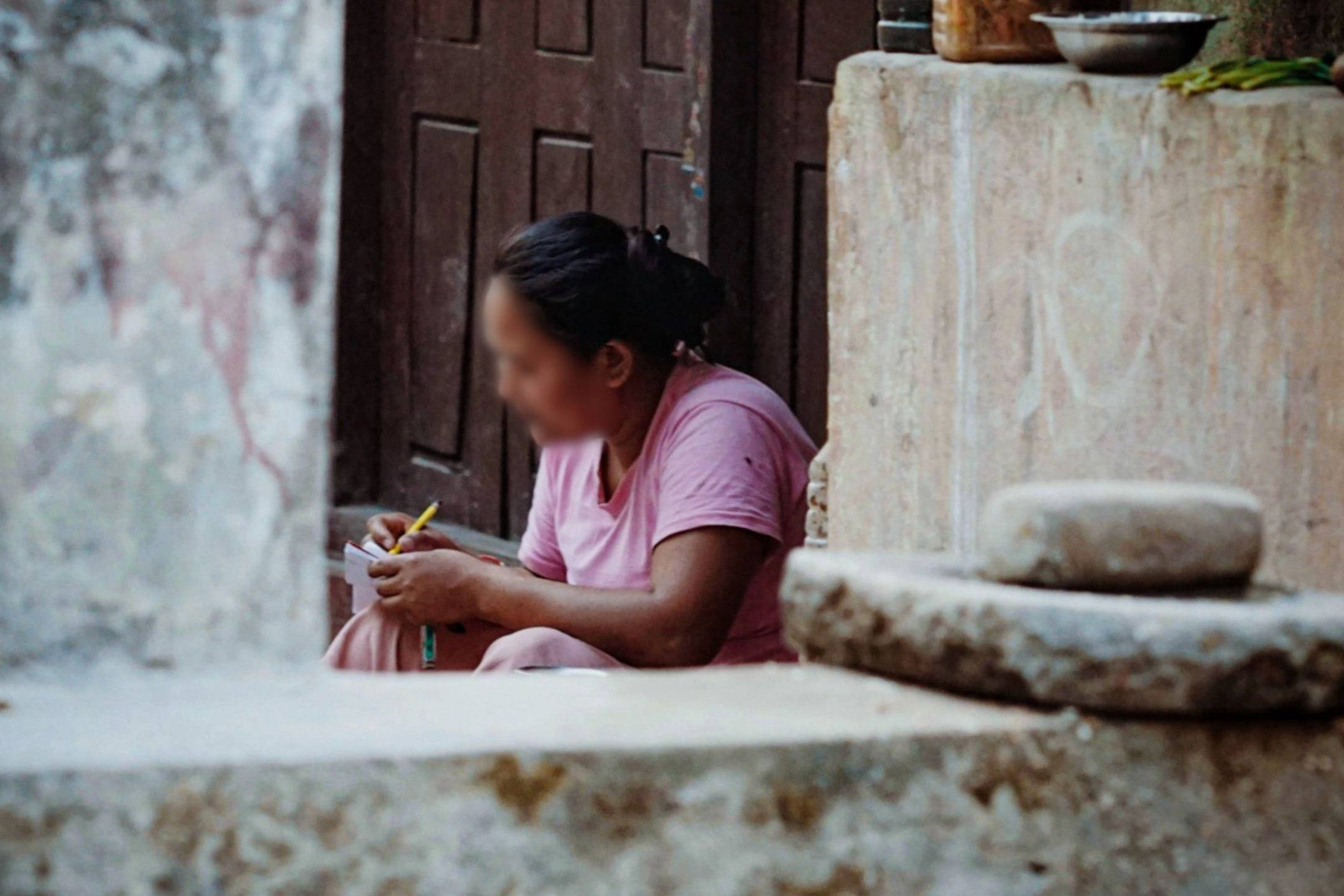 A woman in a pink sari sits on a stone step, writing in a notebook. The scene conveys focus and simplicity, with rustic surroundings including a stone wall.