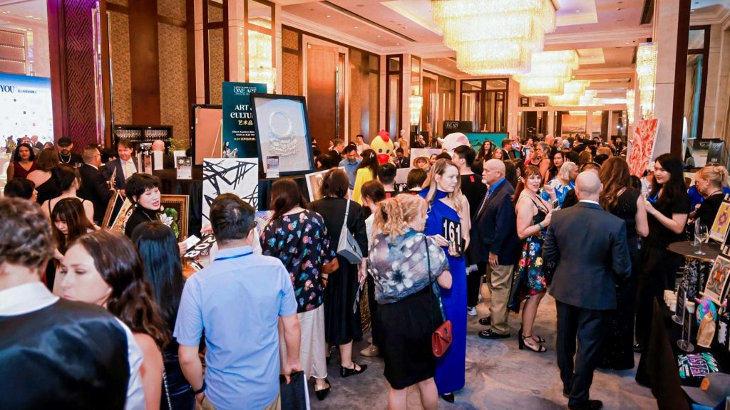 A bustling art exhibition with people mingling, viewing artworks, and chatting. The room is elegantly lit with chandeliers, creating a lively and sophisticated atmosphere.