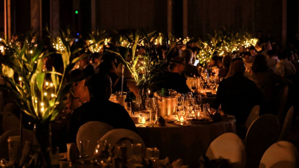 A warmly lit banquet hall with guests seated at round tables, holding small lit candles. The atmosphere is celebratory and elegant, with smiles and soft lighting.