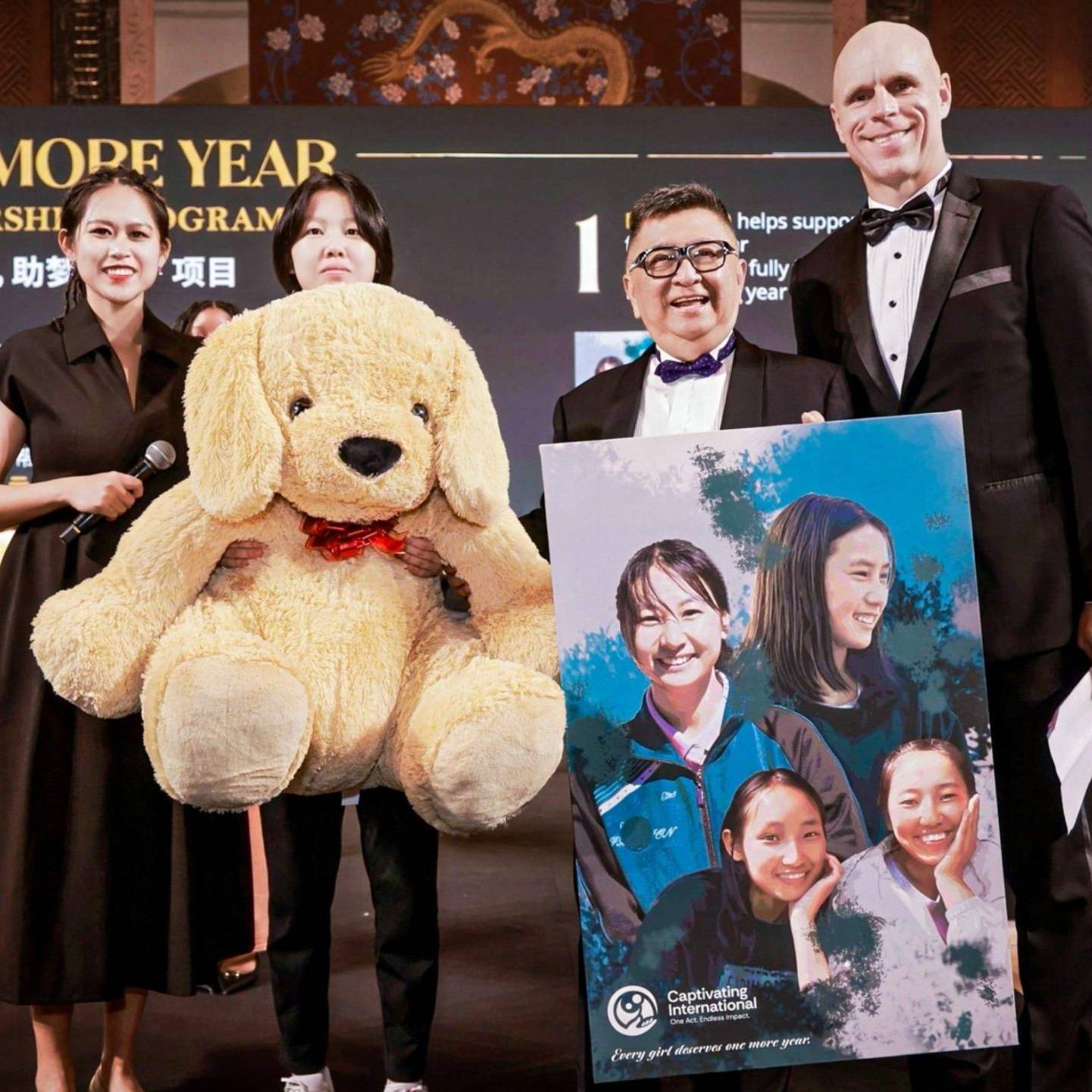 A group stands on stage during a charity event, holding a giant teddy bear and a poster featuring smiling people. The atmosphere is festive and formal.