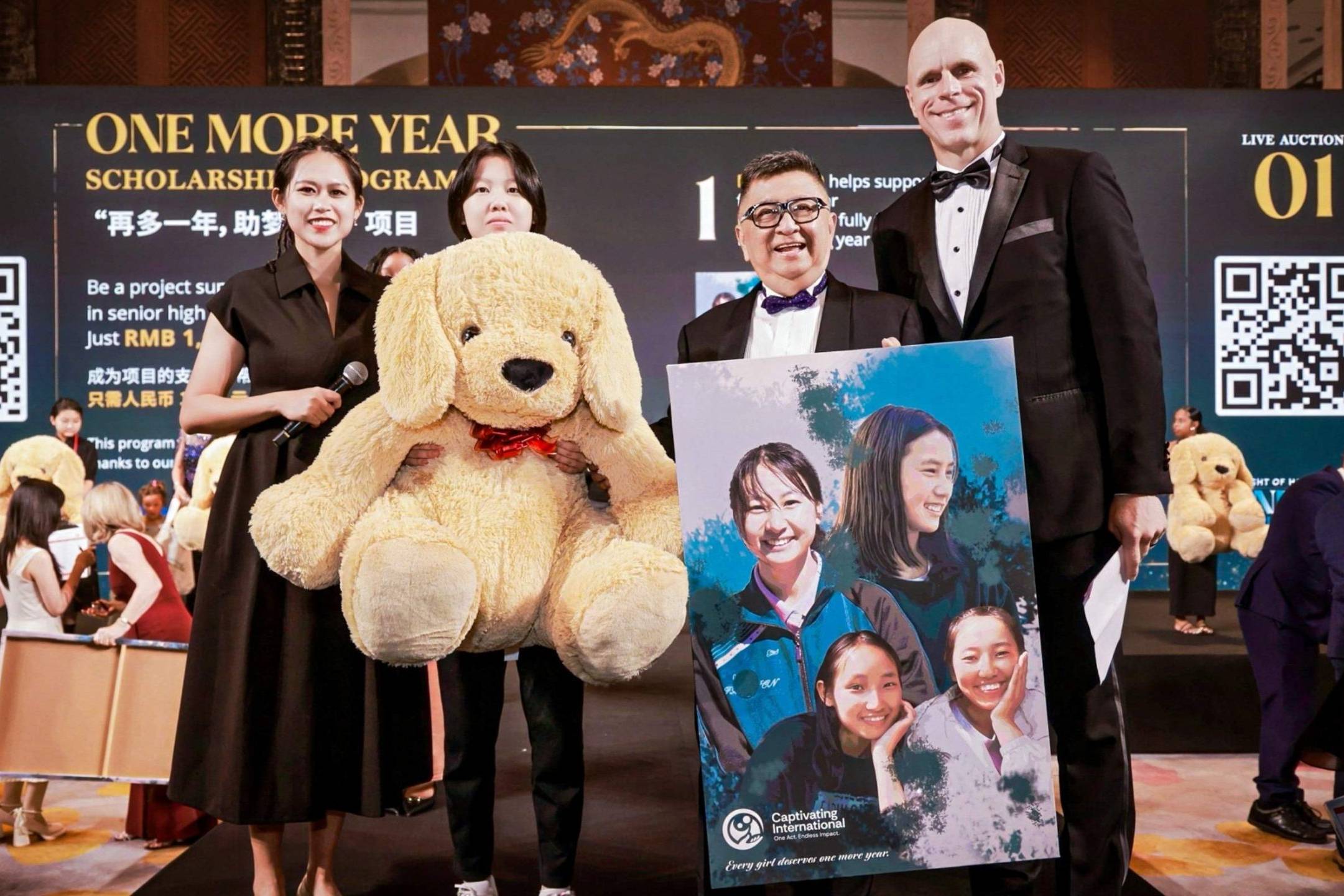 A group stands on stage during a charity event, holding a giant teddy bear and a poster featuring smiling people. The atmosphere is festive and formal.