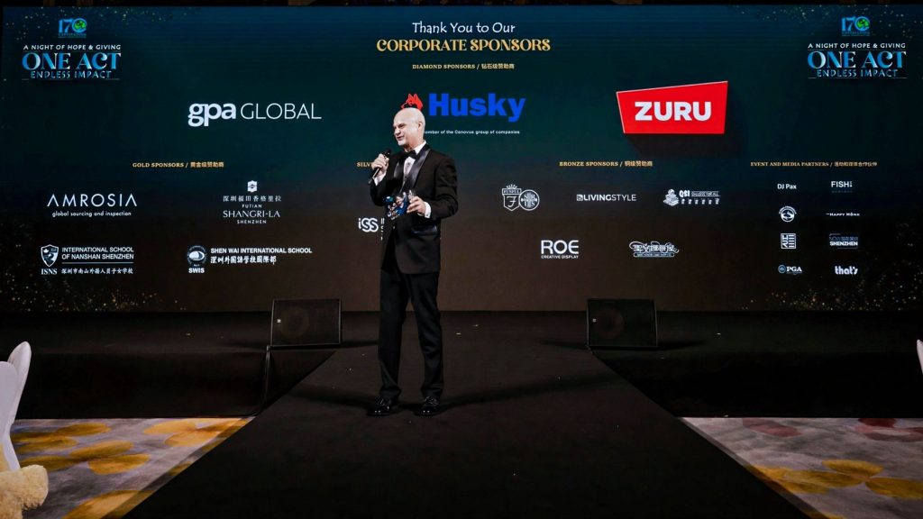 A man in a suit stands on a stage, holding a microphone, in front of a large screen displaying corporate sponsor logos. The setting is formal and professional.