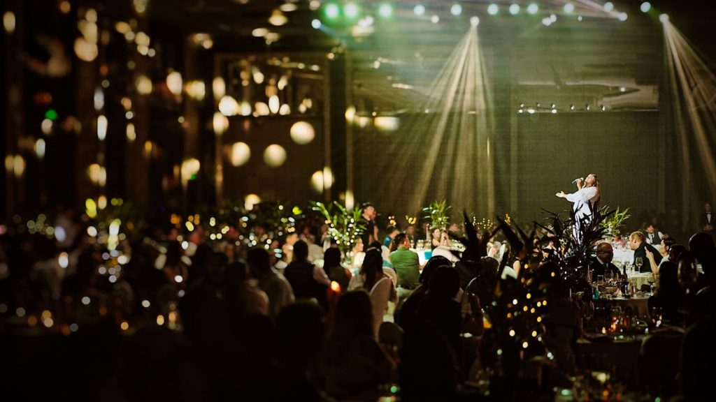 A performer sings under spotlights on a dimly lit ballroom floor, surrounded by seated guests and soft, warm ambient lighting.