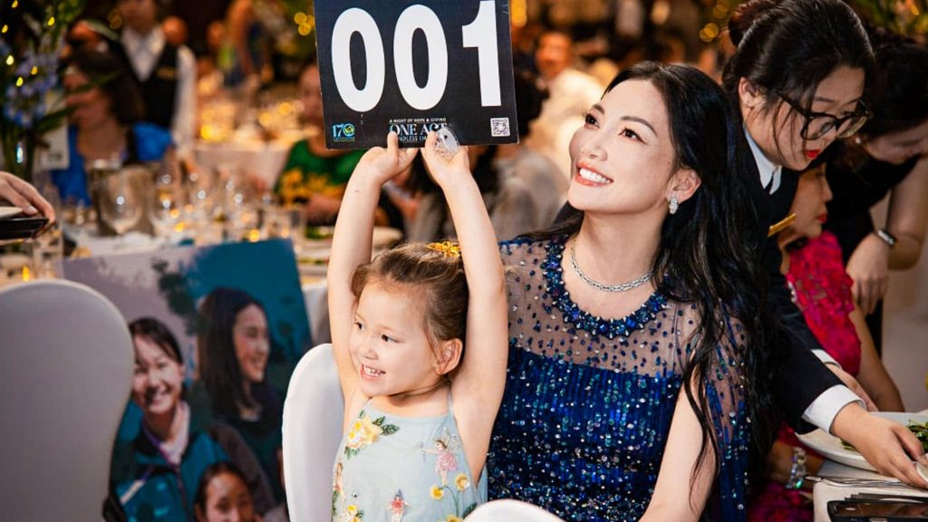 A woman in a blue dress and a young girl excitedly hold up an auction paddle numbered "001" at a formal event. The atmosphere is lively and joyful.