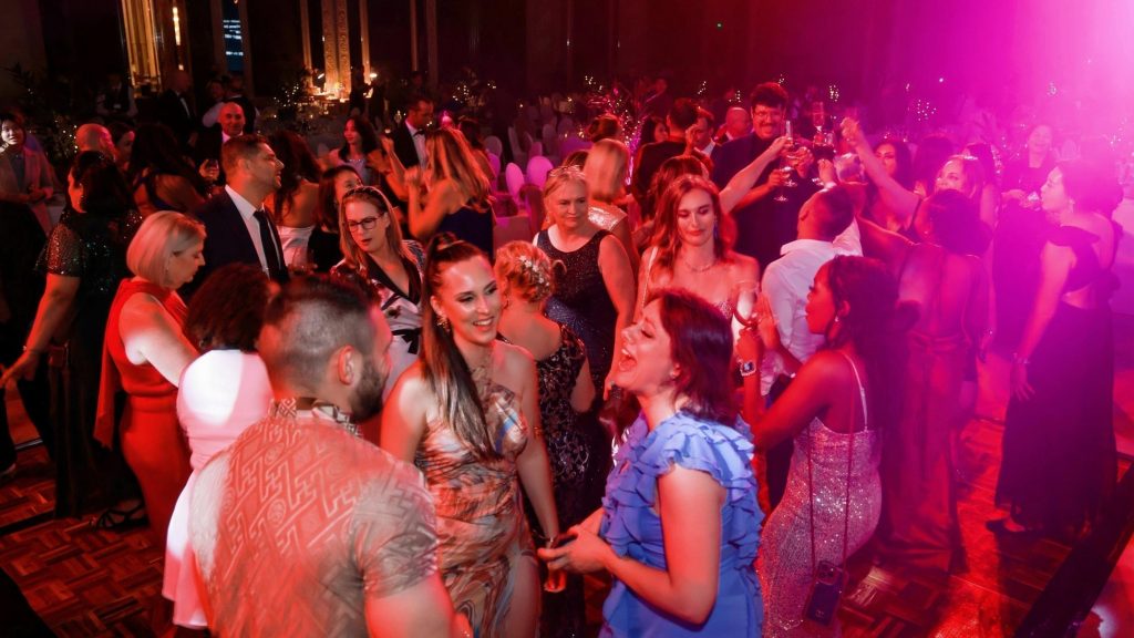 A lively party scene with elegantly dressed people dancing on a dimly lit dance floor, illuminated by vibrant pink lighting, creating an energetic atmosphere.