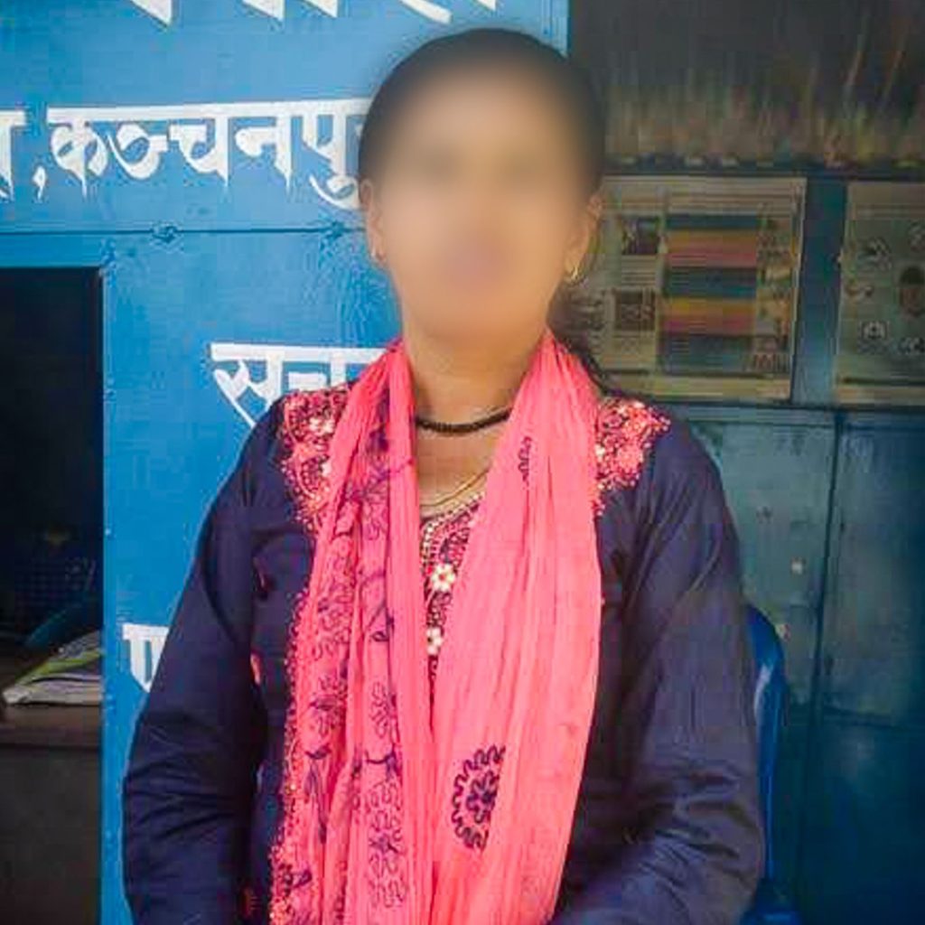 A person standing in front of a blue wall with text, wearing a dark blue outfit and pink scarf. Face blurred for privacy.