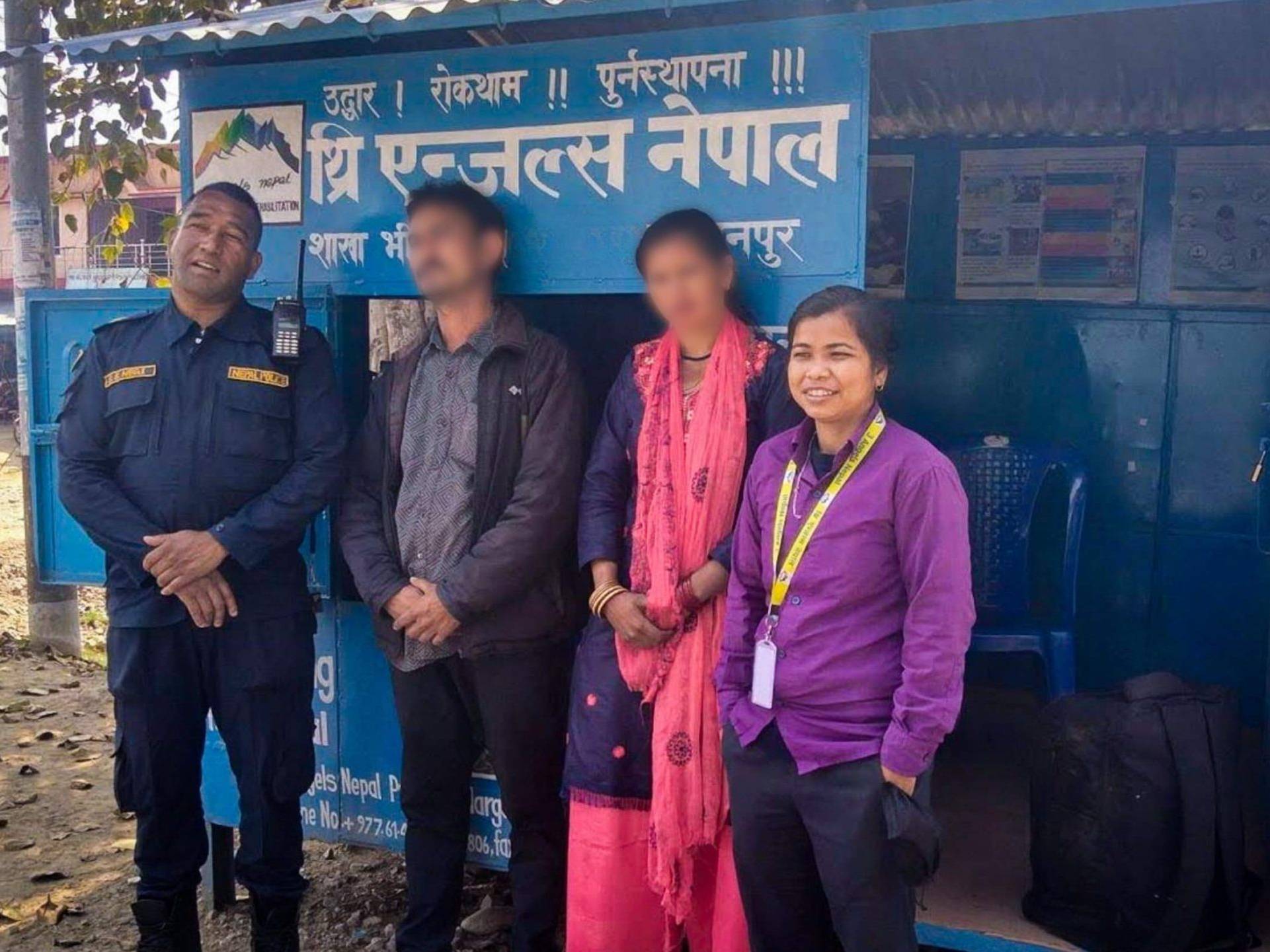 A group of four people are standing in front of a blue roadside stall with white text. One person is in a uniform, and another wears a bright pink outfit. A woman in purple stands beside them smiling. The mood is friendly and relaxed. The faces of the man and woman in the middle are blurred for privacy.