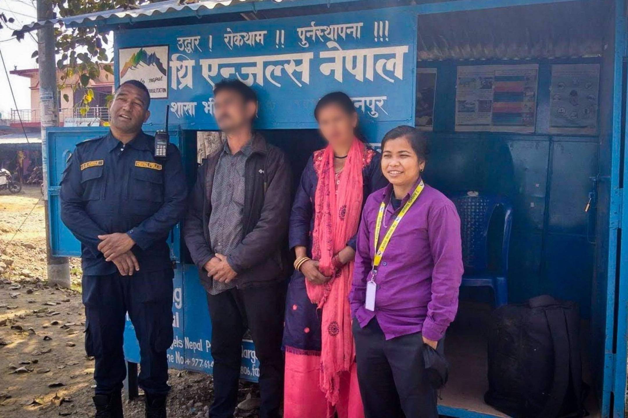 A group of four people are standing in front of a blue roadside stall with white text. One person is in a uniform, and another wears a bright pink outfit. A woman in purple stands beside them smiling. The mood is friendly and relaxed. The faces of the man and woman in the middle are blurred for privacy.