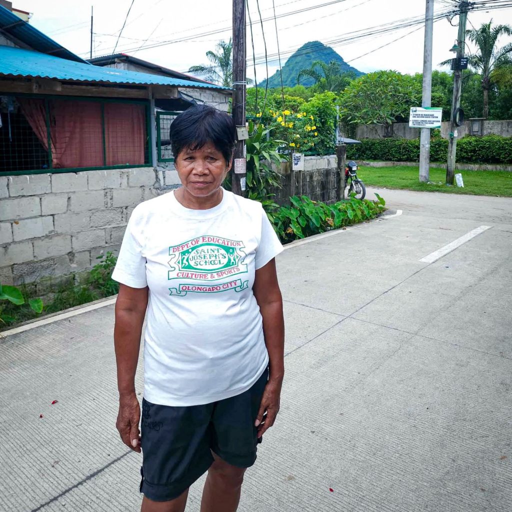 A woman stands on a concrete road in a rural area, wearing a white T-shirt and black shorts. Behind her, a green landscape with a mountain peak is visible. The setting is calm and serene.
