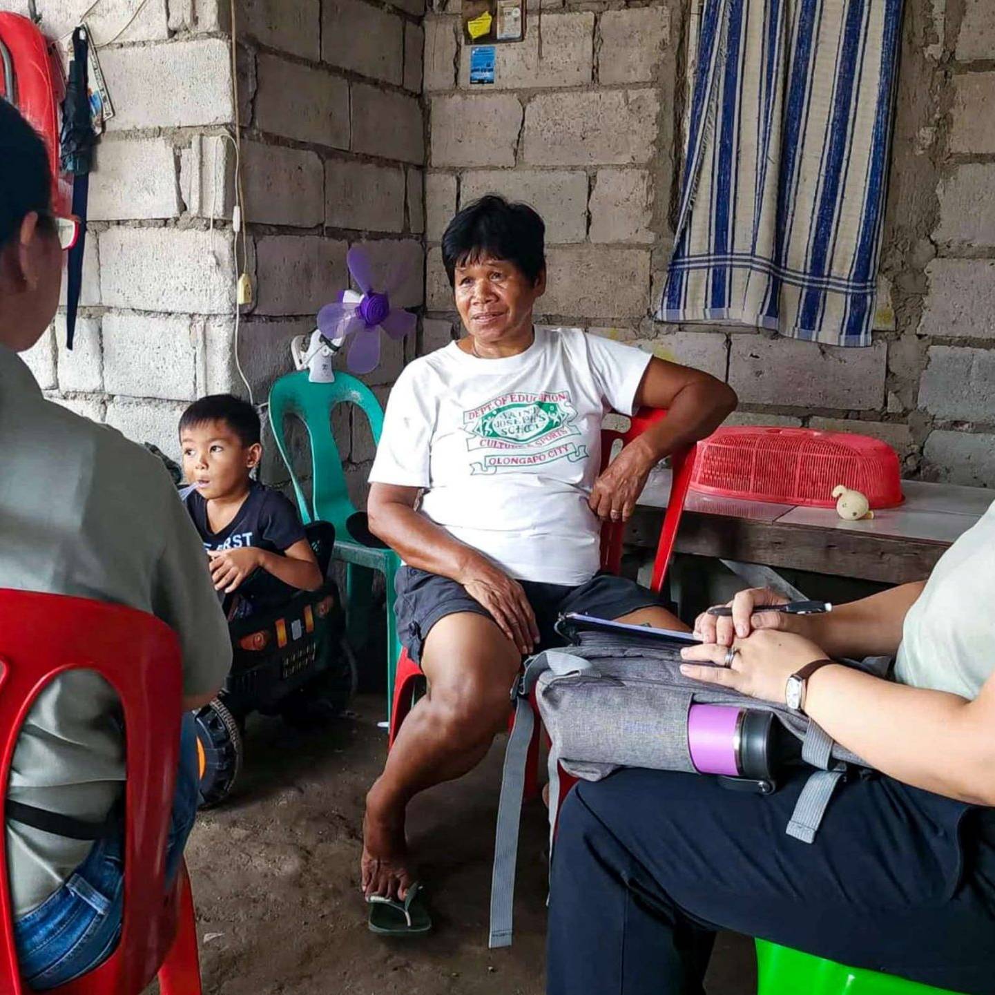 A group of four people, including a child, sit in a modest home with concrete walls. The three adults having a conversation, creating a serious yet calm tone.