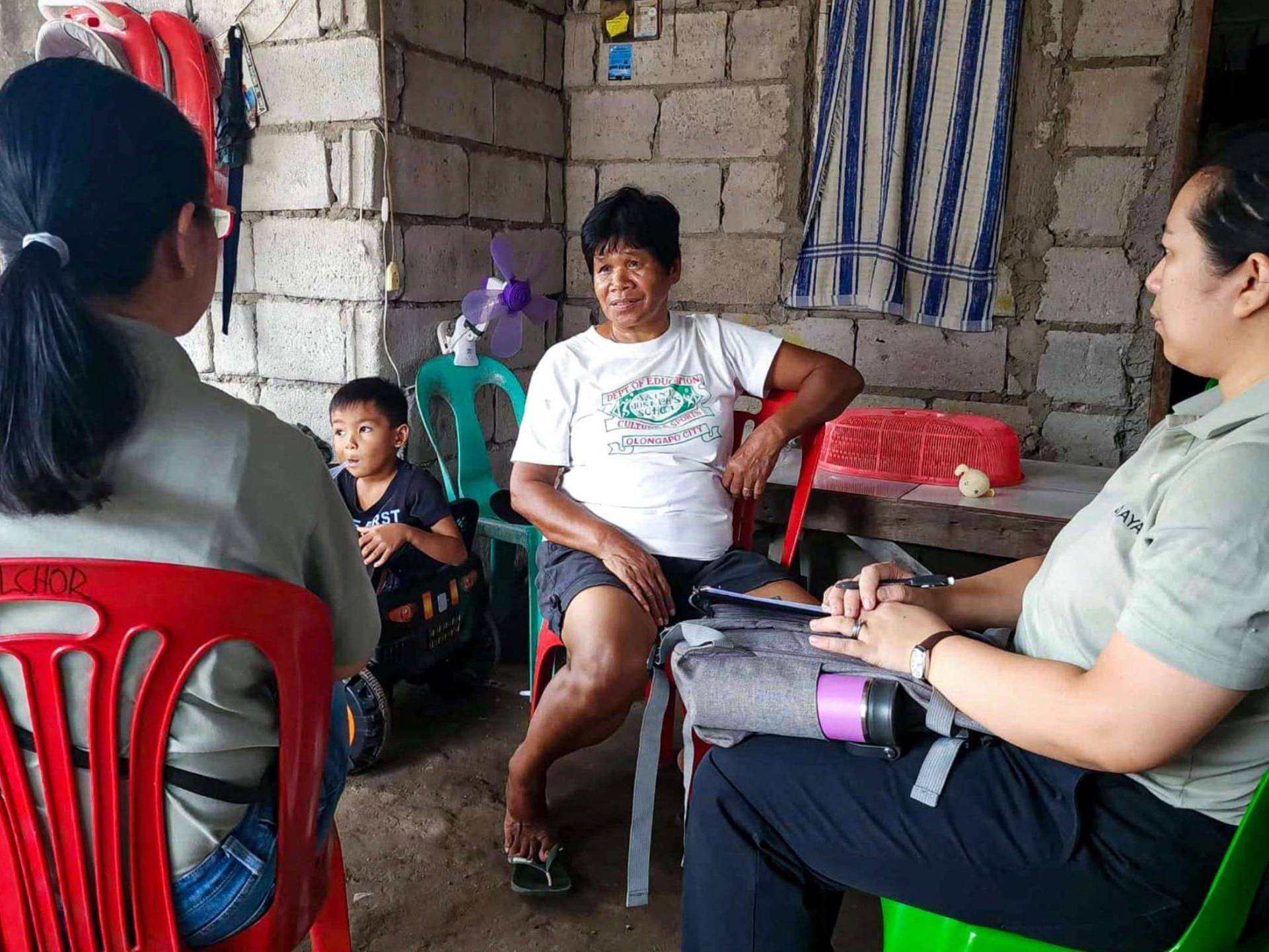 A group of four people, including a child, sit in a modest home with concrete walls. The three adults having a conversation, creating a serious yet calm tone.
