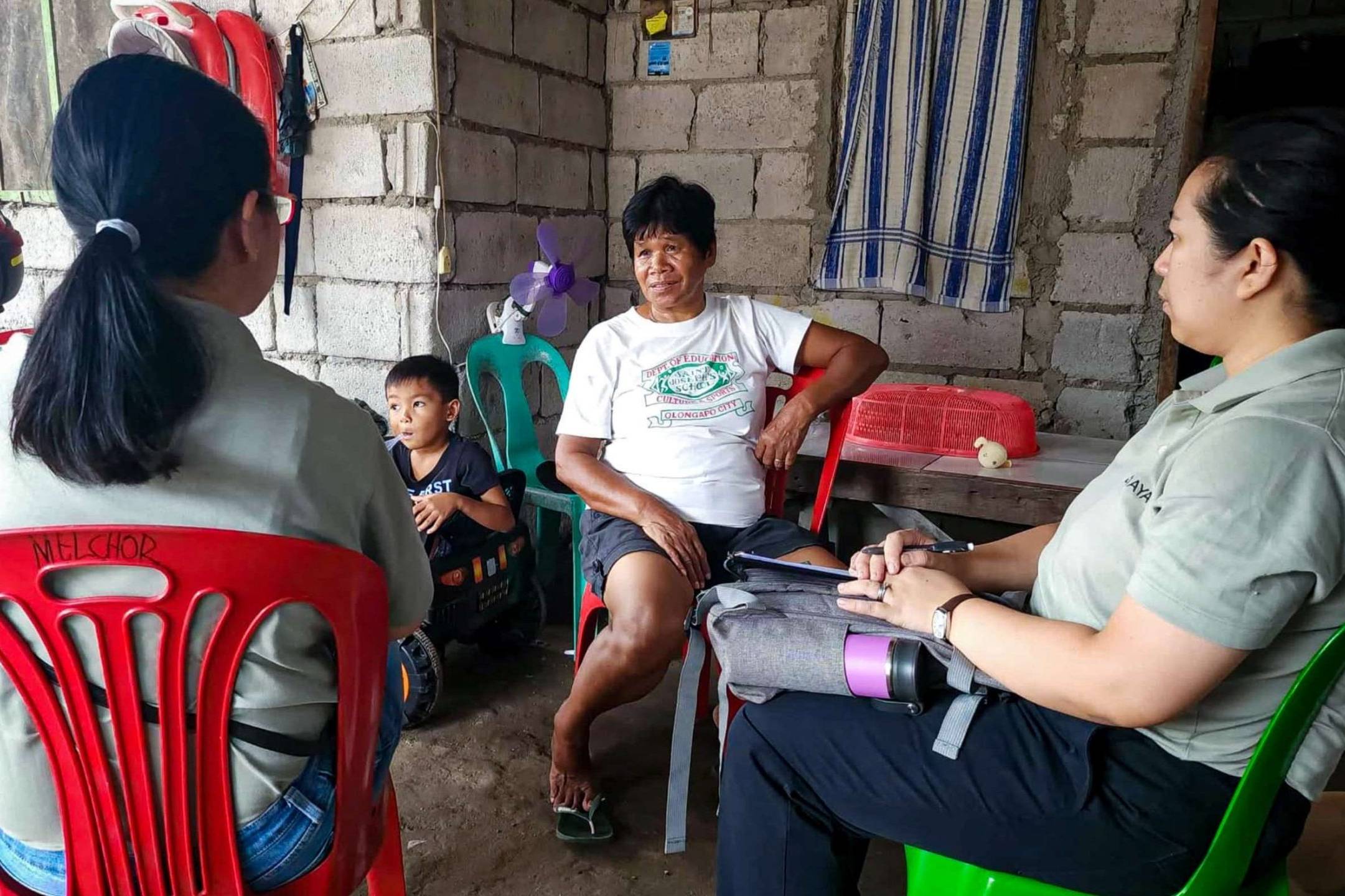 A group of four people, including a child, sit in a modest home with concrete walls. The three adults having a conversation, creating a serious yet calm tone.