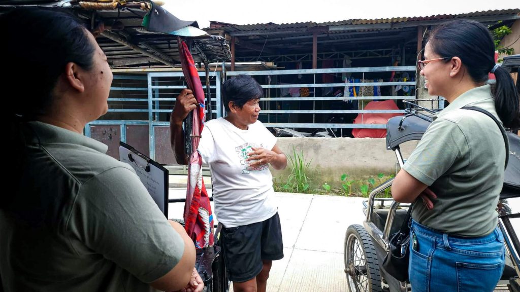 Three people stand outside a rustic building. The central figure gestures expressively, wearing a white shirt. The mood is engaged and conversational.