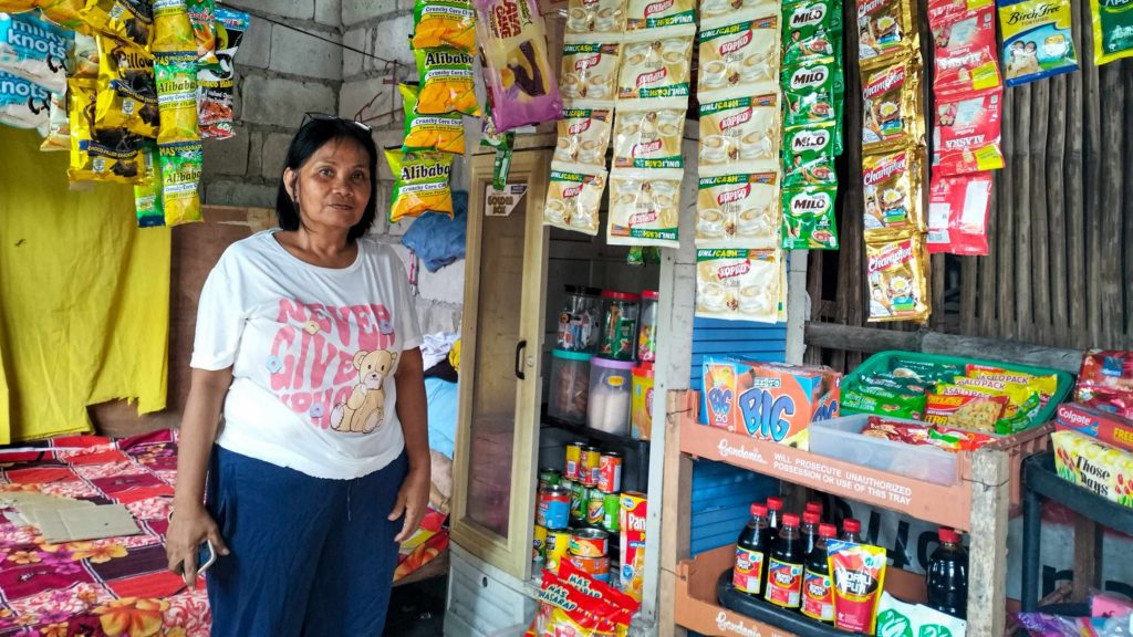 A woman in casual clothes stands in a small, colorful store. Various snacks and drinks are displayed around her, creating a vibrant, lively atmosphere.