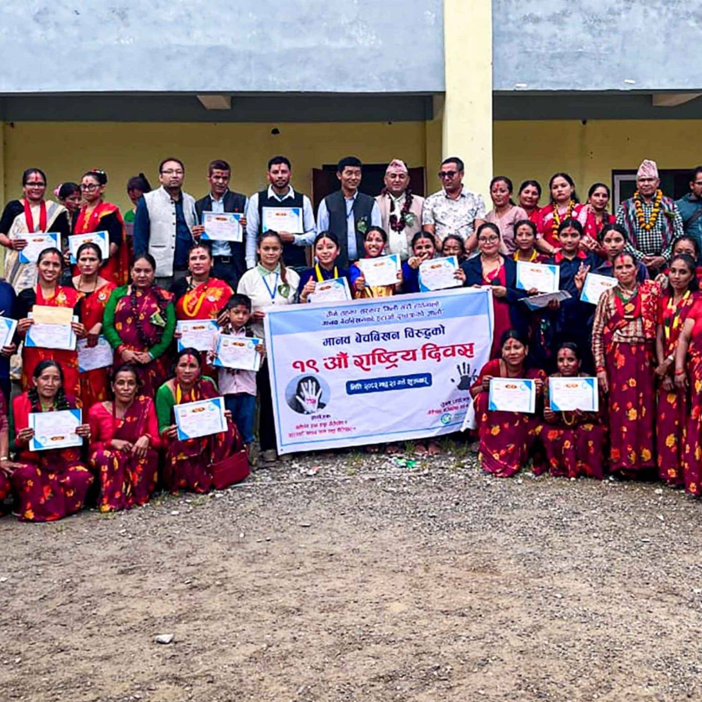 A group of people in colorful traditional attire poses with certificates outside a building. A banner is displayed, creating a celebratory and communal atmosphere.