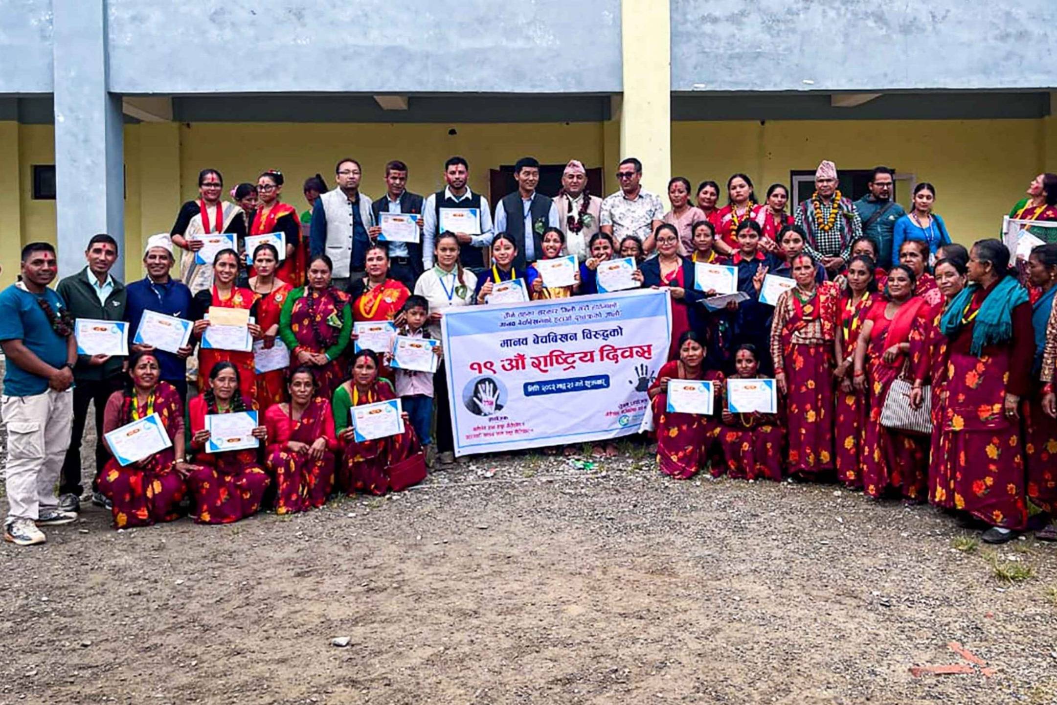 A group of people in colorful traditional attire poses with certificates outside a building. A banner is displayed, creating a celebratory and communal atmosphere.