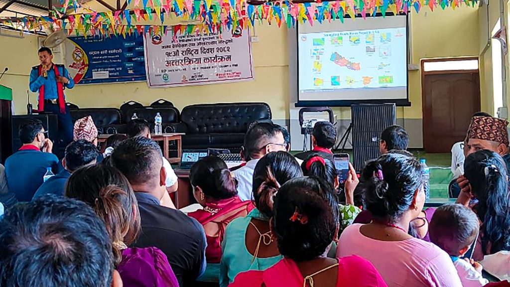 A presenter in a colorful outfit stands by a lectern, gesturing toward a projection screen with maps. Attentive seated audience faces forward, under colorful banners.