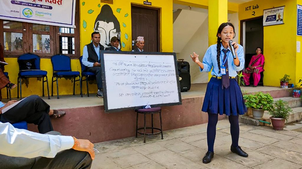 A student in a blue school uniform passionately speaks into a microphone in front of a whiteboard outside a yellow building. Seated adults observe.