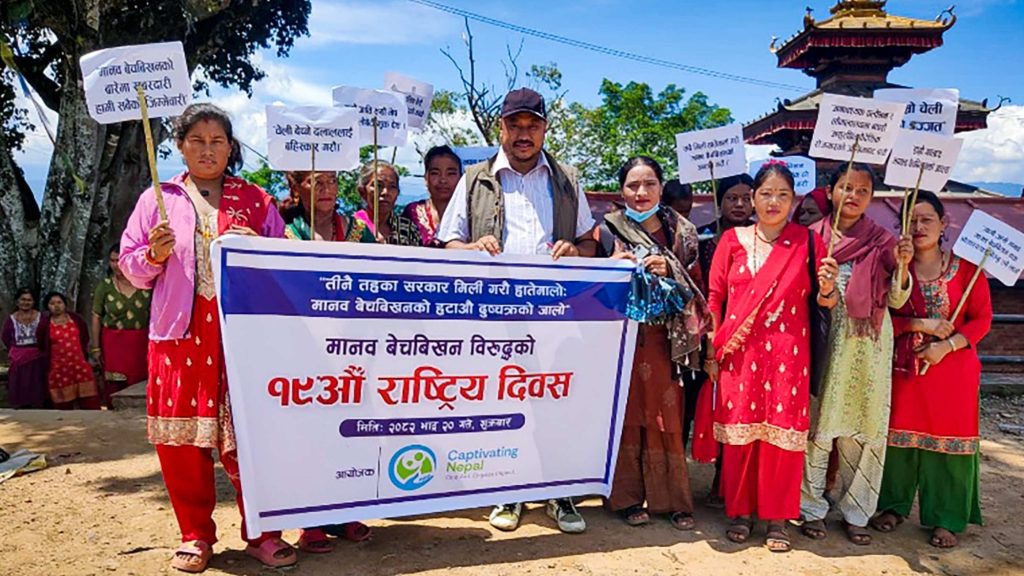 A group of people, mostly women, stand outdoors holding signs and a large banner written in Nepali. They appear to be participating in a protest or awareness event. The women are dressed in traditional clothing, and the mood is determined and unified.