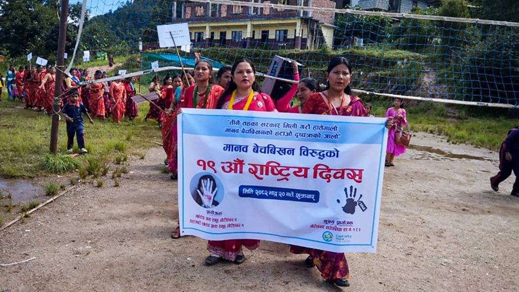A group of women in red traditional attire hold a banner promoting a national day event against human trafficking. The scene conveys unity and activism.