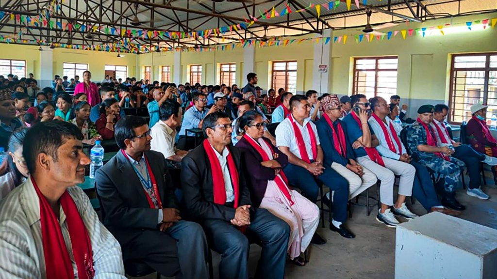 A large, attentive crowd sits in a decorated hall with colorful bunting. People in the front row wear red scarves, suggesting a formal event or ceremony.