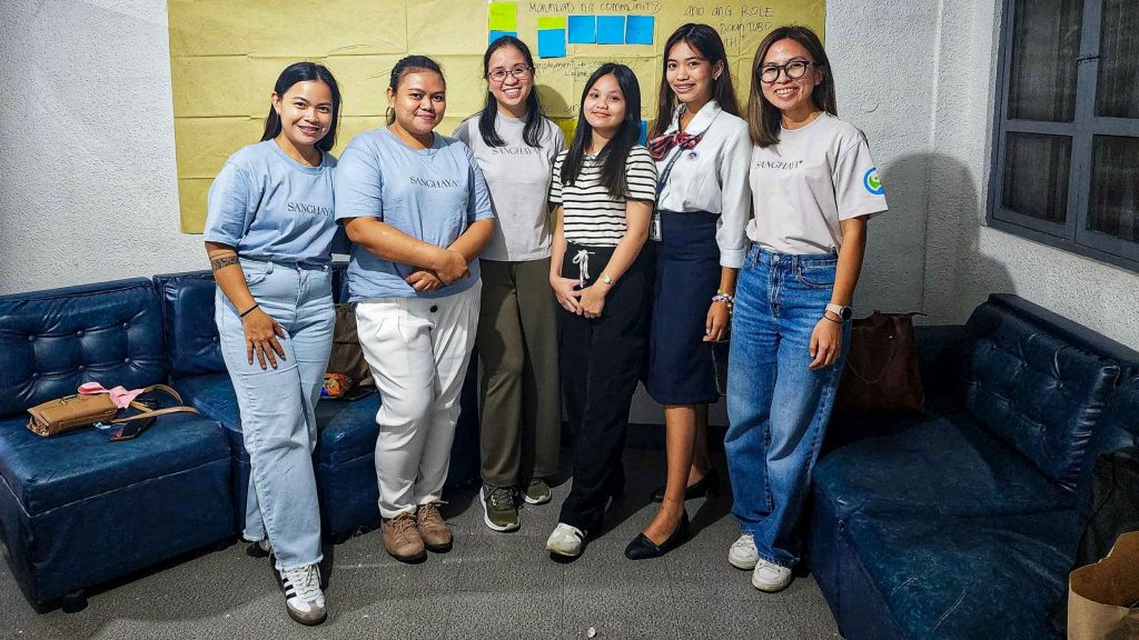 A group of six women pose for a photo in a room with navy blue couches. They are smiling warmly, conveying a friendly and positive atmosphere. Behind them is a yellow wall with colorful sticky notes, suggesting a collaborative or creative setting.