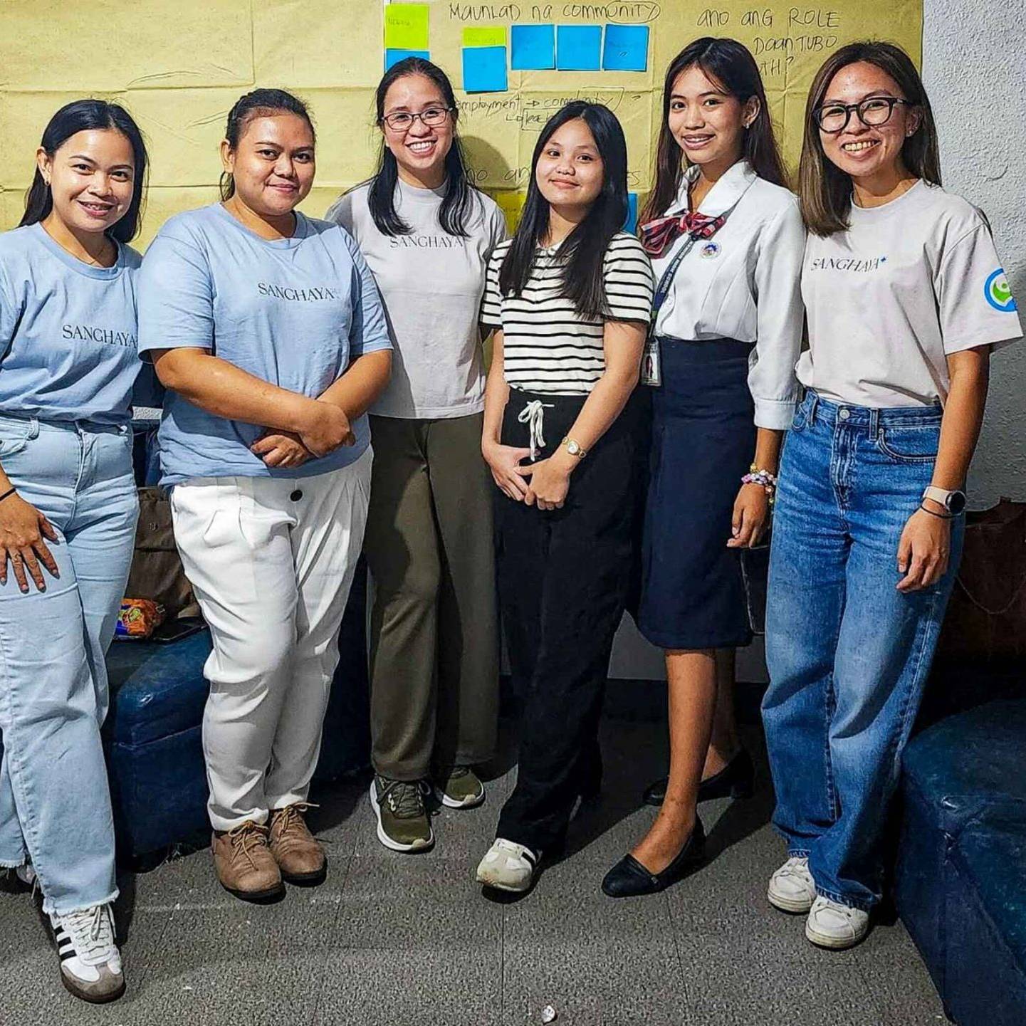 A group of six women pose for a photo in a room with navy blue couches. They are smiling warmly, conveying a friendly and positive atmosphere. Behind them is a yellow wall with colorful sticky notes, suggesting a collaborative or creative setting.