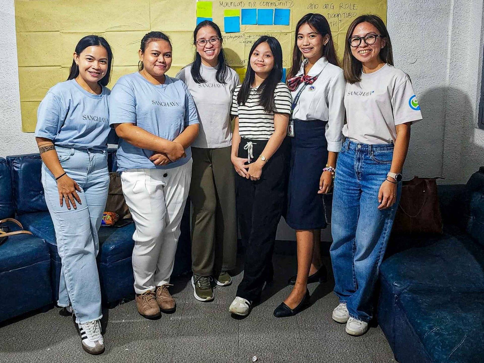 A group of six women pose for a photo in a room with navy blue couches. They are smiling warmly, conveying a friendly and positive atmosphere. Behind them is a yellow wall with colorful sticky notes, suggesting a collaborative or creative setting.