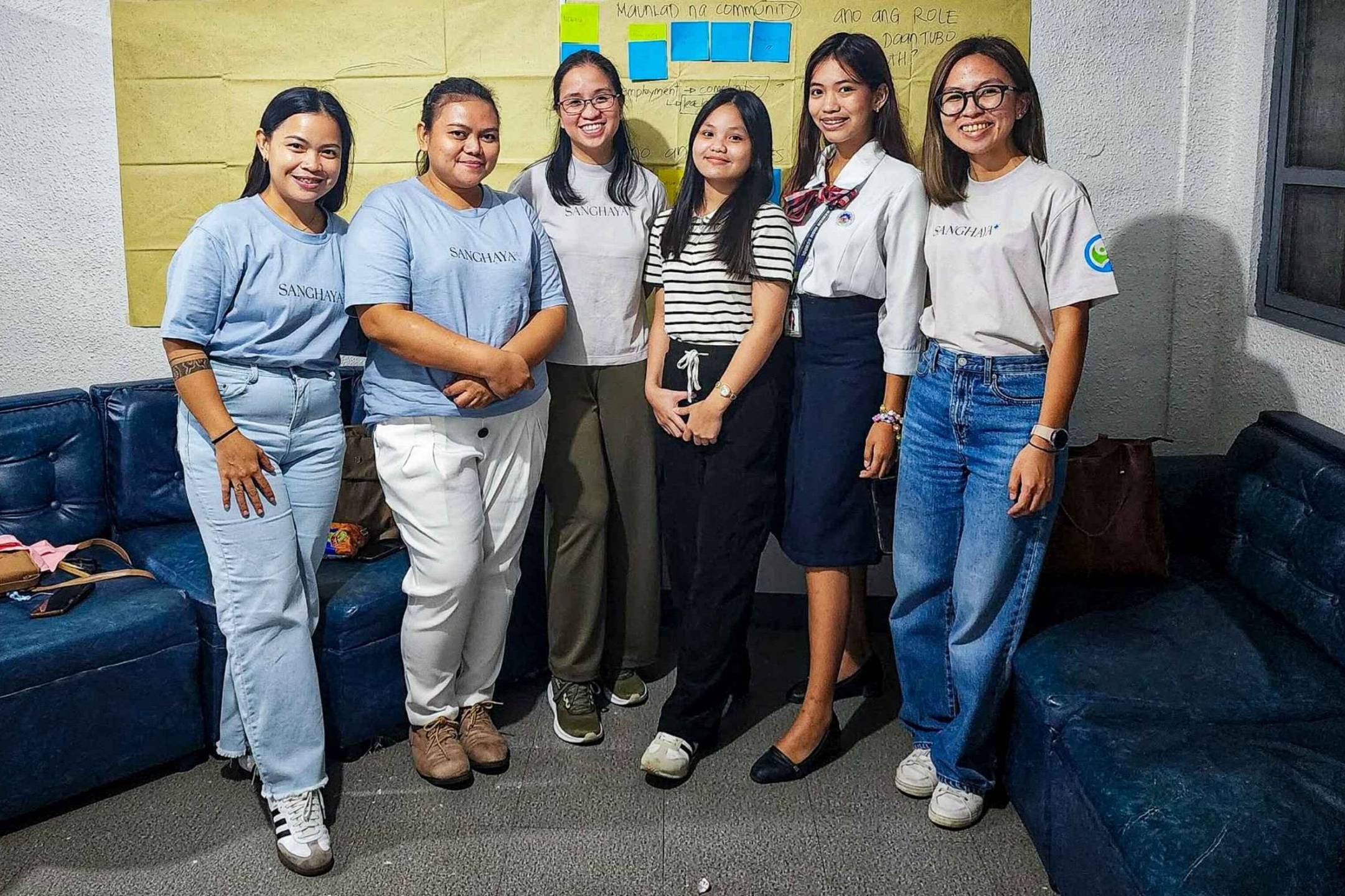 A group of six women pose for a photo in a room with navy blue couches. They are smiling warmly, conveying a friendly and positive atmosphere. Behind them is a yellow wall with colorful sticky notes, suggesting a collaborative or creative setting.