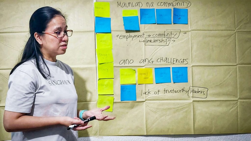 A woman stands in front of a paper-covered wall displaying a workshop board with blue and yellow sticky notes. She gestures while talking, appearing engaged.