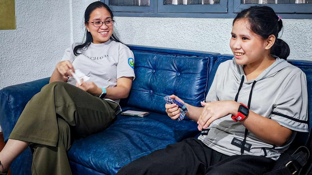 Two women sit on a blue couch, smiling and talking. One holds a small object, and they both wear casual attire, conveying a relaxed, friendly atmosphere.