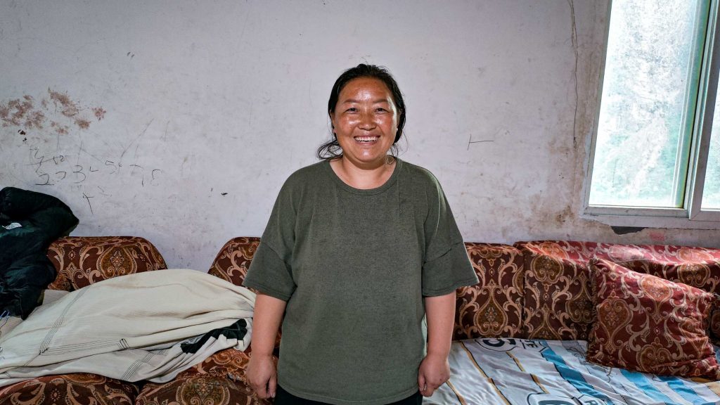 A smiling woman stands in a simple room with a patterned sofa and a window. The atmosphere is warm and inviting, conveying a sense of genuine happiness.
