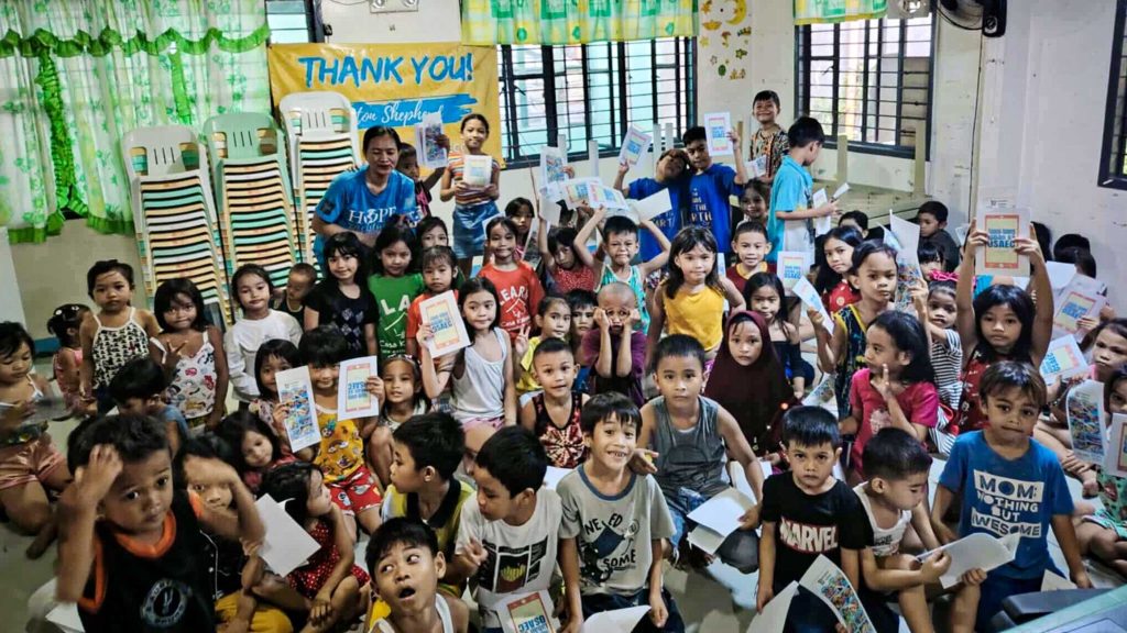 A large group of children in a classroom holds booklets, smiling. The scene is colorful and lively, conveying joy and gratitude.