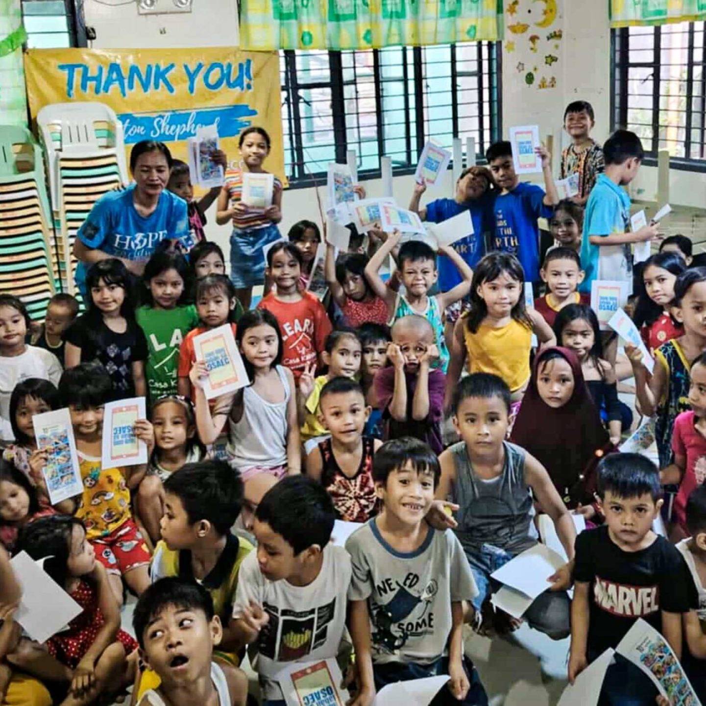 A large group of children in a classroom holds booklets, smiling. The scene is colorful and lively, conveying joy and gratitude.
