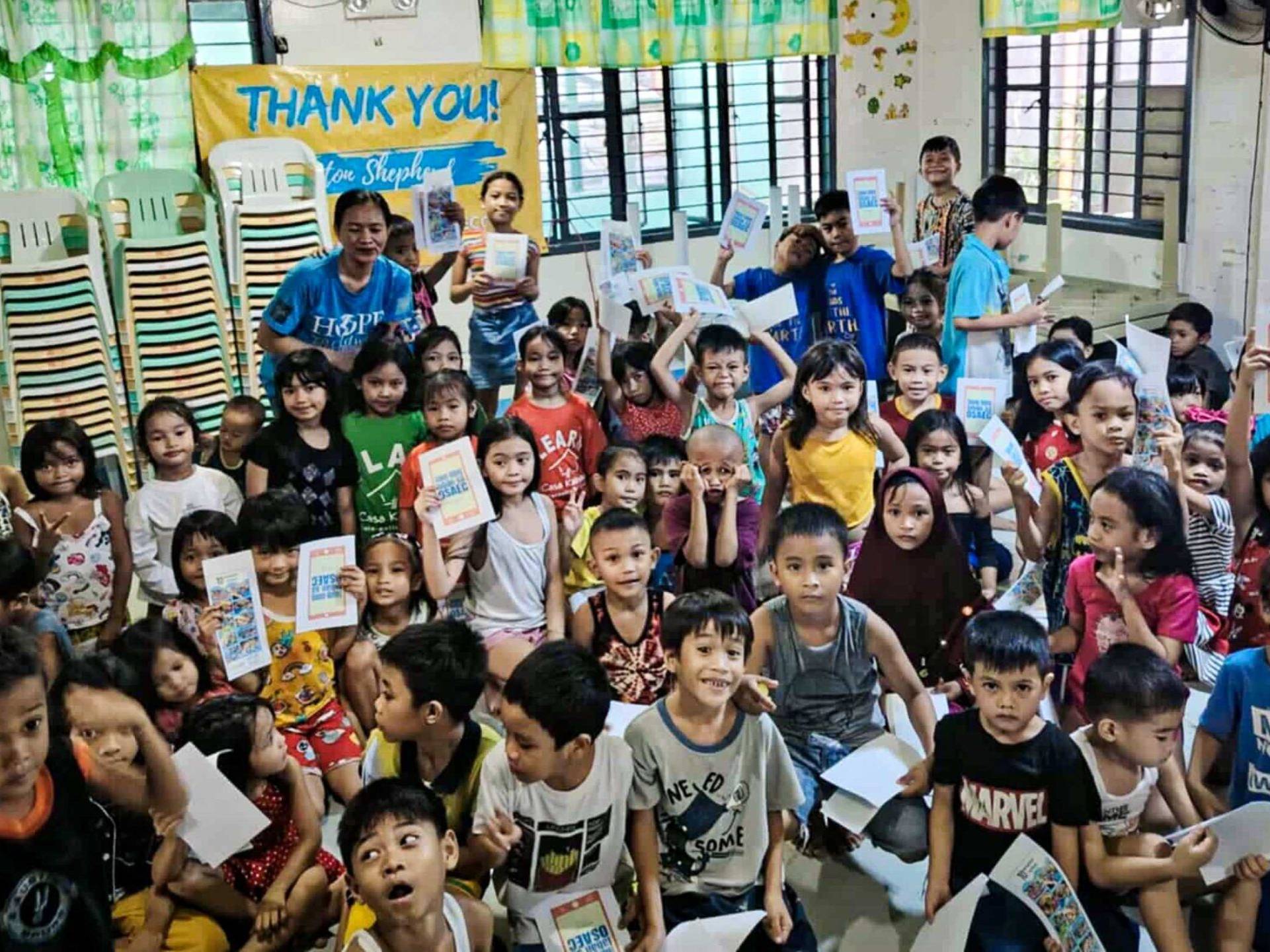 A large group of children in a classroom holds booklets, smiling. The scene is colorful and lively, conveying joy and gratitude.