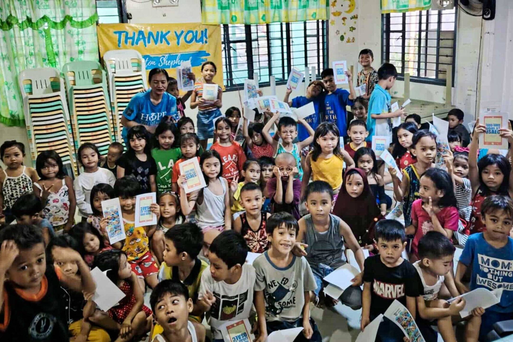 A large group of children in a classroom holds booklets, smiling. The scene is colorful and lively, conveying joy and gratitude.